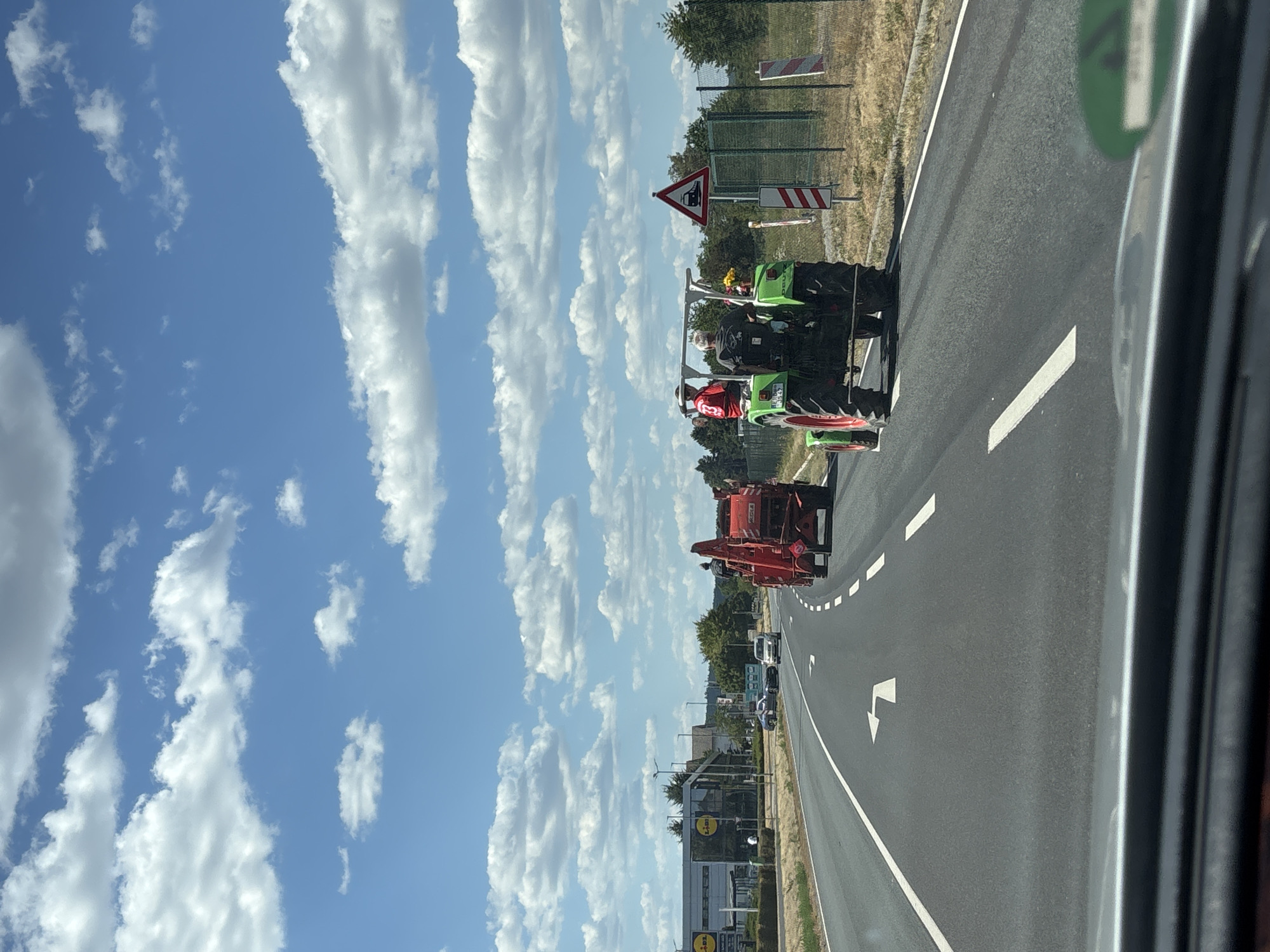 In Wörth am Main, Germany, a procession of agricultural machinery moves along a paved road under a bright blue sky with scattered white clouds. In the foreground, a green tractor with red wheels is visible, carrying two individuals. The driver wears a dark shirt, and a passenger in a red shirt sits beside them. Ahead of the tractor is a red agricultural implement, also with a person visible on top. The road features white lane markings and a turn arrow. To the right of the road, a green fence borders a grassy area, and a triangular sign with a train symbol is posted. In the background, a row of trees and buildings, including a Lidl supermarket, line the side of the road. The perspective suggests the image is taken from a vehicle on the road, observing the procession.