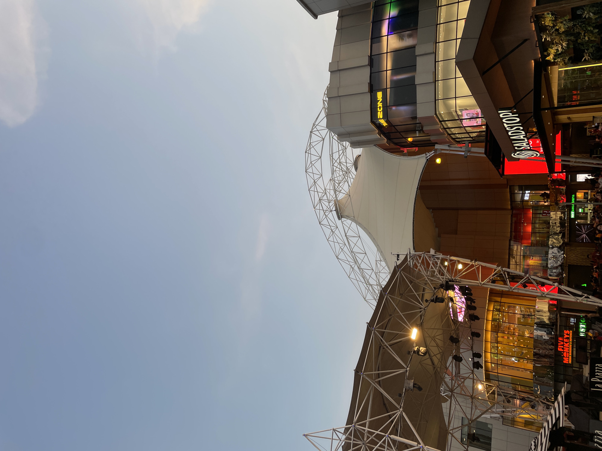 The image depicts an outdoor scene in North Jakarta, Indonesia, likely at or near the Mall of Indonesia. The view is angled upwards, showcasing architectural elements and commercial signage. Visible structures include a large tensile structure or awning supported by a white steel framework, along with adjacent buildings with retail spaces. Businesses with lighted signage such as "SALADSTOP!", "ZONE", "FIVE MONKEYS FRESH BURGERS & HOTDOGS" and "La Piazza" are identifiable. The sky is clear and light blue. The scene suggests a commercial or entertainment area within the city, possibly during dusk or late afternoon.