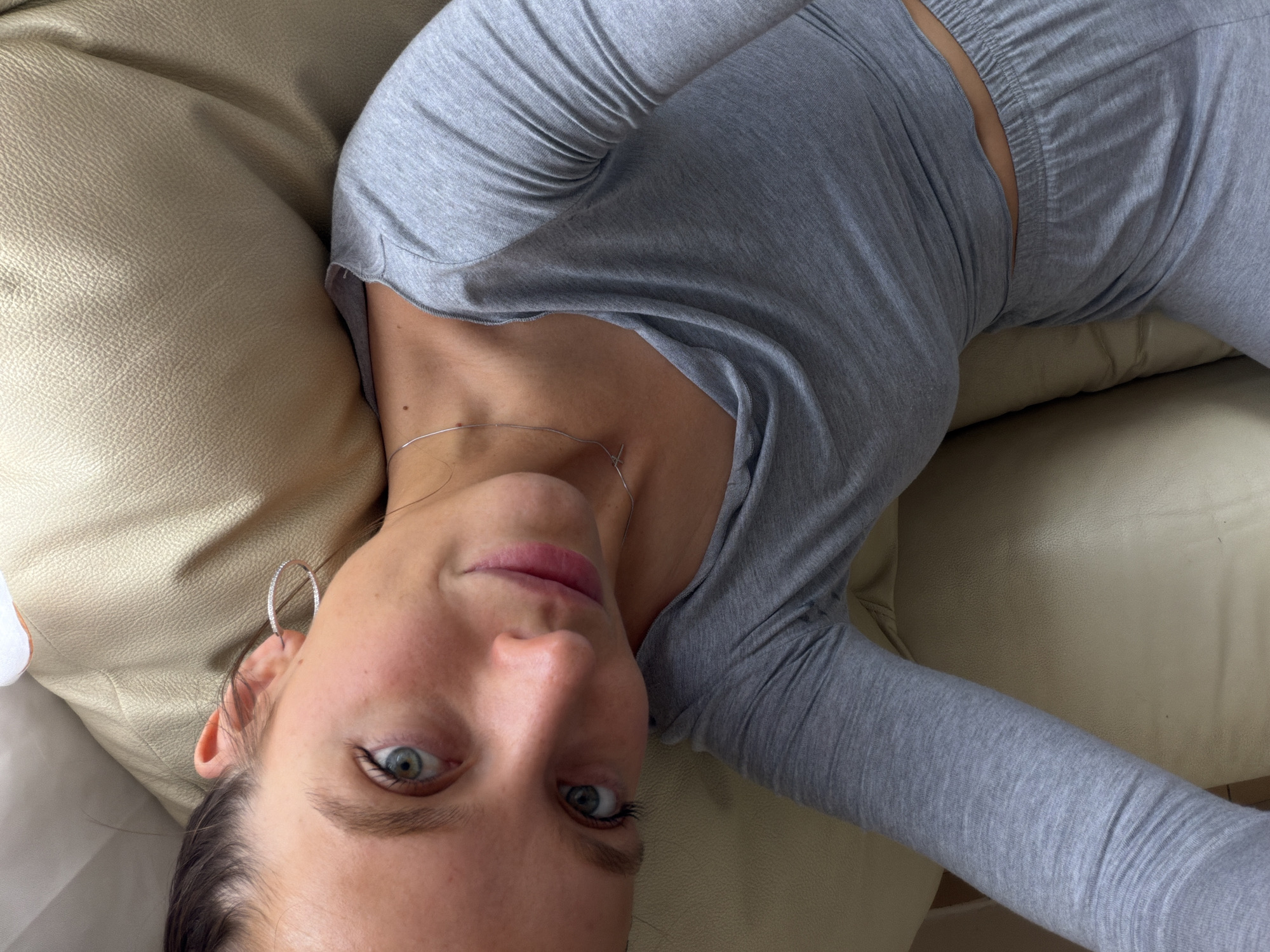 An upside-down, close-up selfie shows a young adult female lounging on a light-colored, possibly leather or faux leather, sofa or cushion. She is looking directly at the camera with her head inverted, her eyes open, and a subtle, relaxed smile on her face. Her eyes are light-colored (blue-green). She is wearing a simple, light grey long-sleeved top and matching grey sweatpants/loungewear bottoms. A thin silver-colored necklace with a small, delicate pendant rests on her collarbone, and she wears a large, silver-colored hoop earring, possibly embellished, in her right ear. The lighting is soft and even, suggesting an indoor setting. There is no visible text in the image. The overall mood is casual and relaxed. The location context provided is Budva, Montenegro.