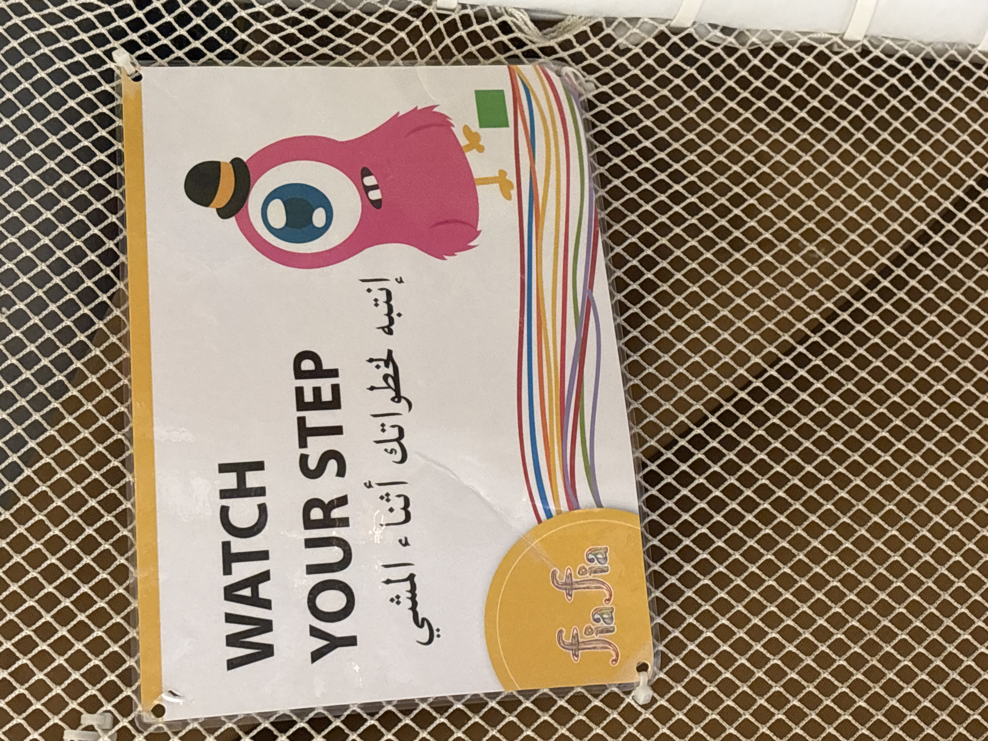 A sign with a cartoon monster and text in English and Arabic is attached to a white mesh fence. The English text reads "WATCH YOUR STEP". The Arabic text translates to "Watch your steps while walking." The monster is pink with one eye and a small hat. A yellow circular logo with the text "fia fia" is in the bottom left corner of the sign. The sign is hanging in what appears to be an indoor play area or children's attraction.