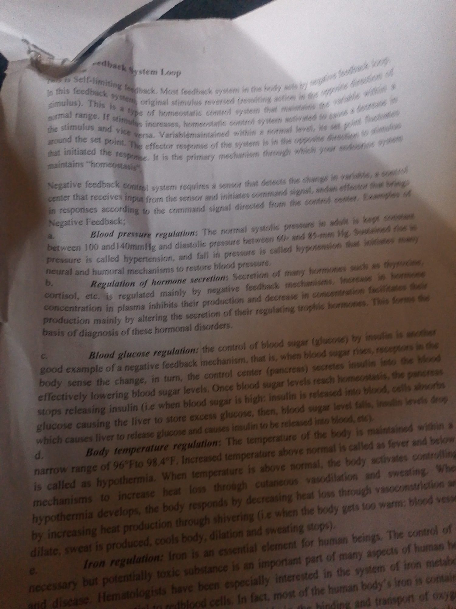 This is a close-up, overhead shot of a printed page from a textbook. The page is filled with dense black text, printed on white paper. The text discusses biological concepts related to feedback systems, specifically focusing on negative feedback mechanisms in the human body. Topics covered include self-limiting feedback, blood pressure regulation, hormone secretion, blood glucose regulation, body temperature regulation, and iron regulation. The image is slightly blurry, suggesting it may have been taken with a mobile phone camera. The lighting appears to be artificial, possibly from an overhead room light, as there are no discernible shadows or natural light cues. The focus is entirely on the text, with no other objects or subjects visible in the frame. The context provided indicates the location is Jalingo, Nigeria, but there are no visual cues within the image itself to support this.