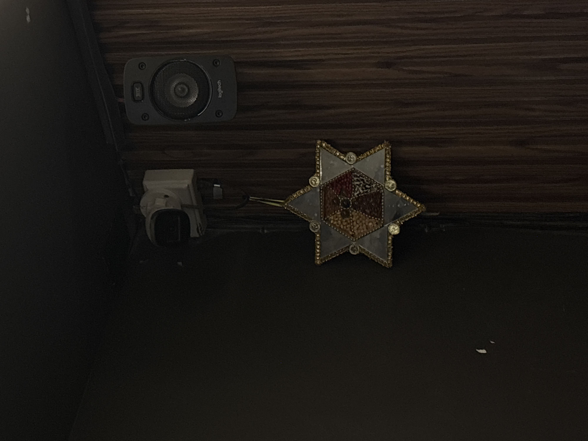 The image presents a dimly lit, low-angle perspective of what appears to be an interior space. The wall is covered with dark, horizontally grained wood paneling. Mounted on the wall is a black Logitech speaker, positioned near a white power adapter with a plug connected to an electrical outlet. Adjacent to the power adapter is an ornate, star-shaped decorative object. It features intricate detailing, possibly made of glass and metal, with a design of alternating geometric patterns and a metallic-gold trim. The lower section of the image reveals a dark, undefined surface, possibly a wall or floor, marked by a couple of faint, white flecks. The overall environment suggests an indoor setting, potentially a room in a house. The scene is devoid of people and actions, focusing instead on the arrangement of the objects and the ambient darkness. The location is specified as Puebla, Mexico; however, there is no definitive evidence within the image to confirm this geographical setting, so it is only relevant context.