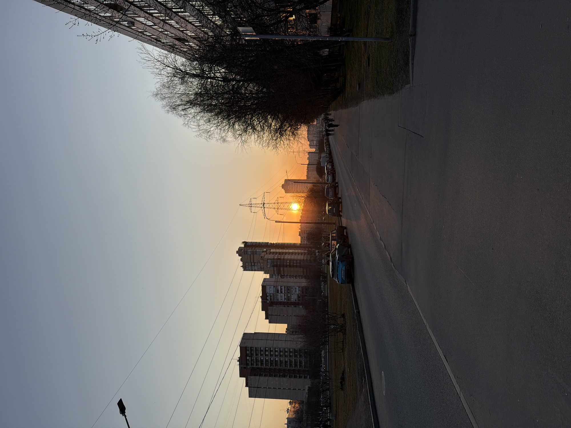 The photo depicts a street in Saint Petersburg, Russia, at sunset. Tall apartment buildings line the left side of the road, with bare trees and power lines silhouetted against the setting sun. Several cars are parked along the right side of the street. People are walking in the distance. The sky is light blue transitioning to orange near the horizon. The street surface appears to be asphalt.