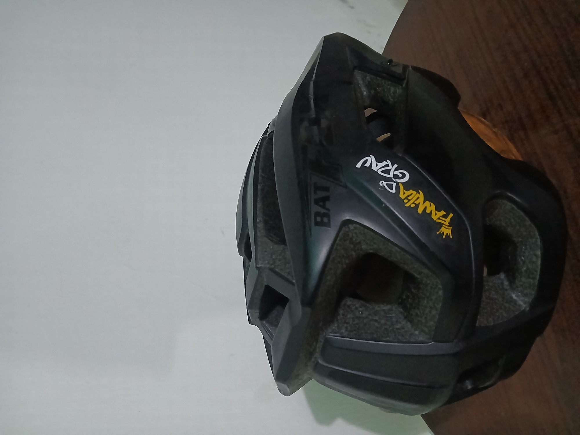 A black bicycle helmet is positioned on a dark brown wooden surface, with a white wall visible in the background. The helmet has the word "BAT" printed vertically on its side, as well as the words "Familia Grau" and a crown symbol in yellow. The exact location is not discernable from the image.