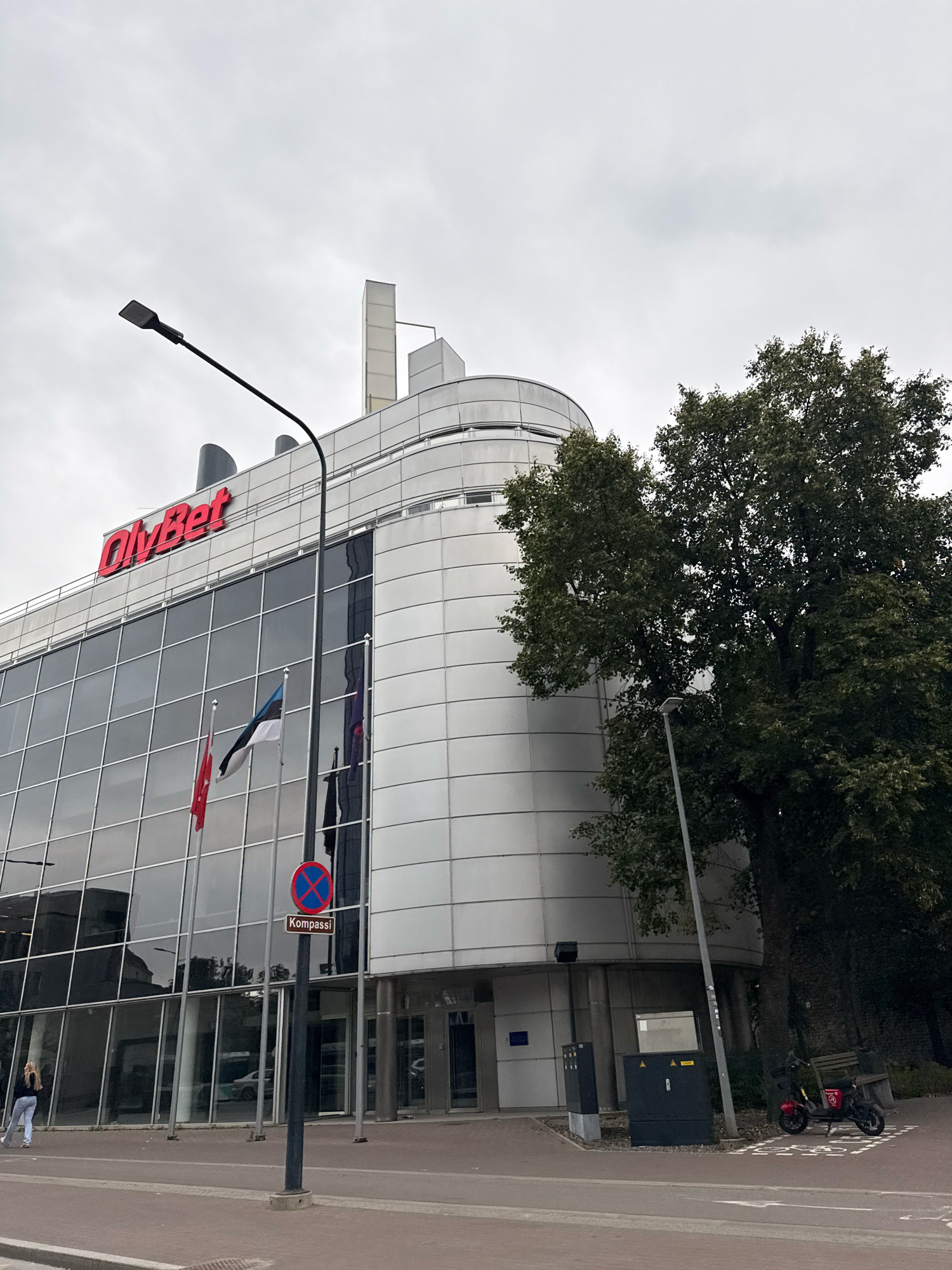 The image captures the OlyBet building, a modern structure with a glass facade and silver metallic paneling, located in Tallinn, Estonia. The building features a prominent red "OlyBet" logo at the top. Two flags, one Estonian and one red, are displayed on flagpoles in front of the building. A street lamp stands to the right of the entrance, and a traffic sign indicating "Kompassi" and a no-parking symbol is visible on a pole. In the foreground, a red electric scooter is parked in a designated area. A large, mature tree with dense foliage is situated to the right of the building, casting shadows. The sky is overcast, suggesting a cloudy day. A person in casual attire walks away from the camera on the sidewalk in front of the building.