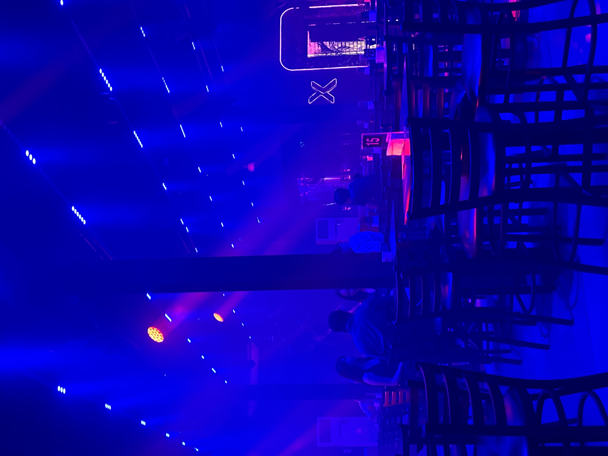 This media file captures a vibrant indoor scene, likely a nightclub or a bar, illuminated with intense blue and purple lighting. The dominant element is the dynamic use of light, with numerous small white lights forming diagonal patterns across the background and larger, circular stage lights casting colorful beams.
The subjects are primarily people, seen in silhouette due to the low lighting and the direction of the light sources. They are seated at tables or standing, suggesting a social gathering or a performance. The chairs themselves, with their dark, angular frames, contribute to the overall atmosphere.
In the background, a prominent neon sign displays a stylized 'X' within a rounded rectangle, suggesting a brand or a venue name. Adjacent to this, a sign with the number "15" is visible, possibly indicating a table number or a zone. The overall scene evokes a sense of energy and excitement, typical of a nightlife setting. The lighting and the presence of people suggest it is nighttime. The location context of Davao City, Philippines, adds a geographical element to this lively urban scene.