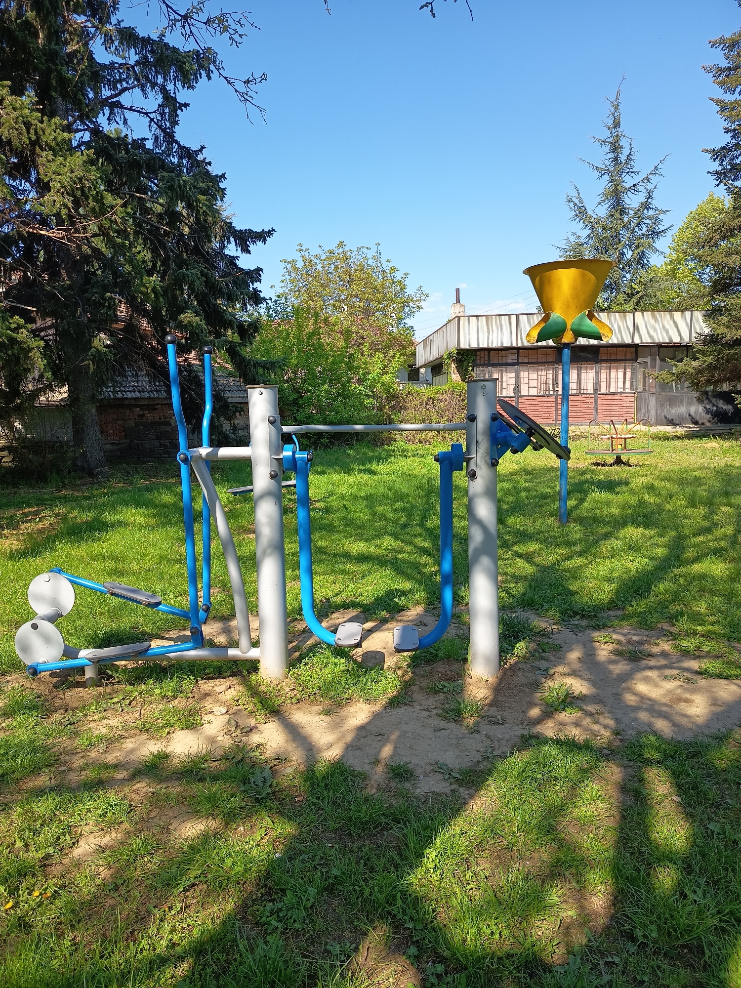 The image captures an outdoor exercise area in an urban environment. An exercise machine painted in silver and blue stands prominently, with stepping platforms and hand grips. Another exercise structure, featuring a yellow funnel-shaped component, is visible behind the first machine. The ground is covered with grass and patches of dirt. Trees surround the area, and a building can be seen in the background. The sky above is clear and blue.