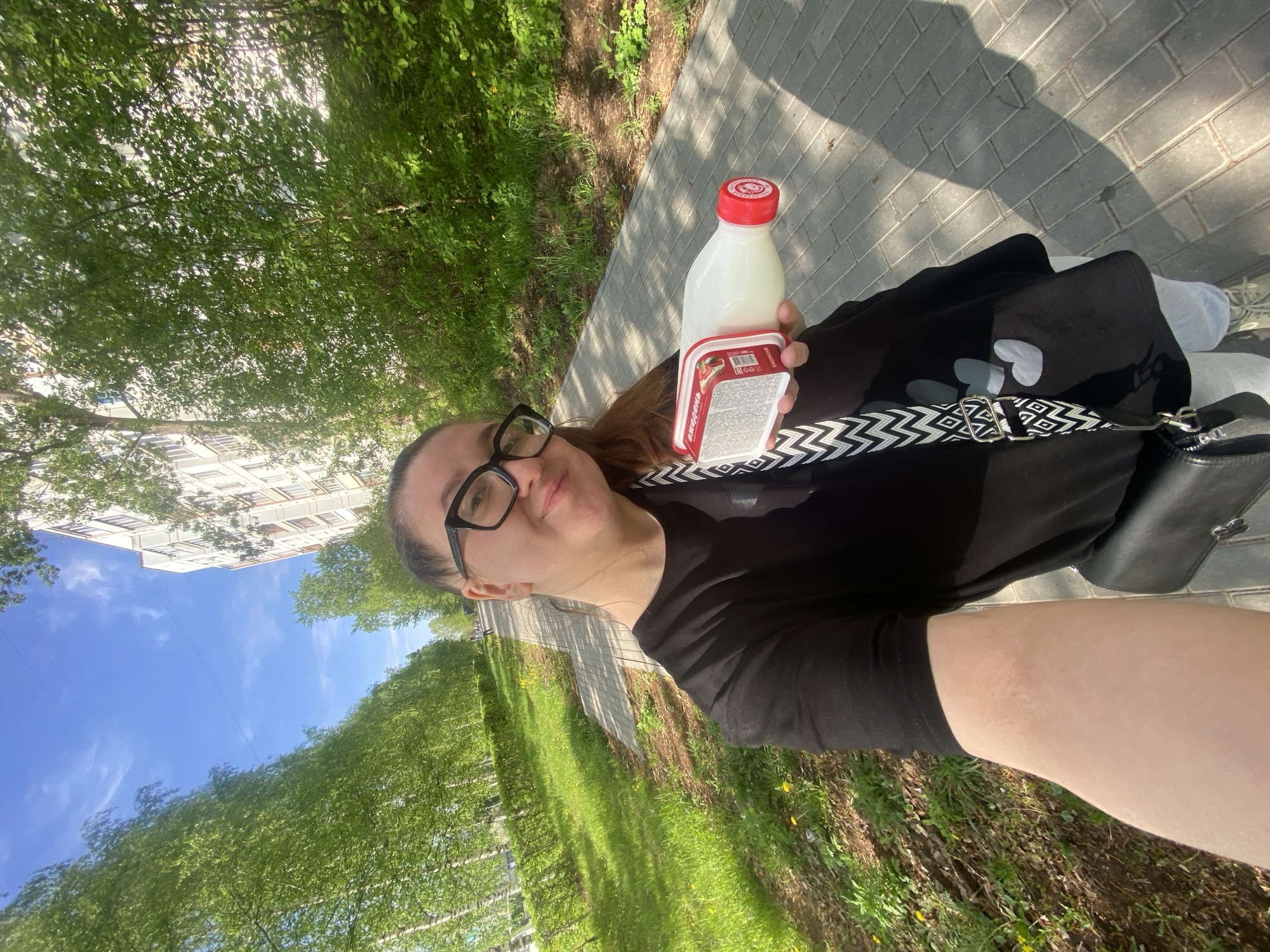 A woman is standing on a sidewalk with brick paving, near a grassy area with trees, potentially in Chaikovsky, Russia. She is holding a bottle of milk with a red cap and a container of what appears to be curd cheese. She is wearing glasses, a black t-shirt with heart designs, and a black bag with a white and black patterned strap. A building is visible in the background. The weather appears to be sunny.
