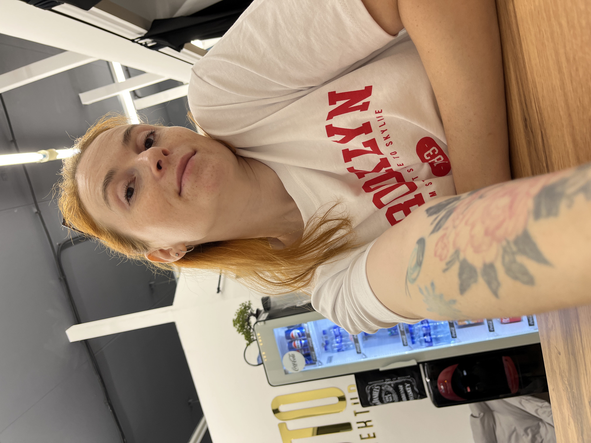 The image shows a young woman with red hair and freckles, wearing a white t-shirt with "BROOKLYN" printed on it in red lettering. She has a tattoo of flowers on her left arm. She is sitting at a wooden table, and in the background, there is a Coca-Cola refrigerator filled with drinks, a black bottle of Jack Daniel's, and a wall with a large golden sign that appears to be a logo or branding. The overall environment suggests an indoor public space, possibly a cafe or a lounge area. The image was taken in Chaikovsky, Russia.