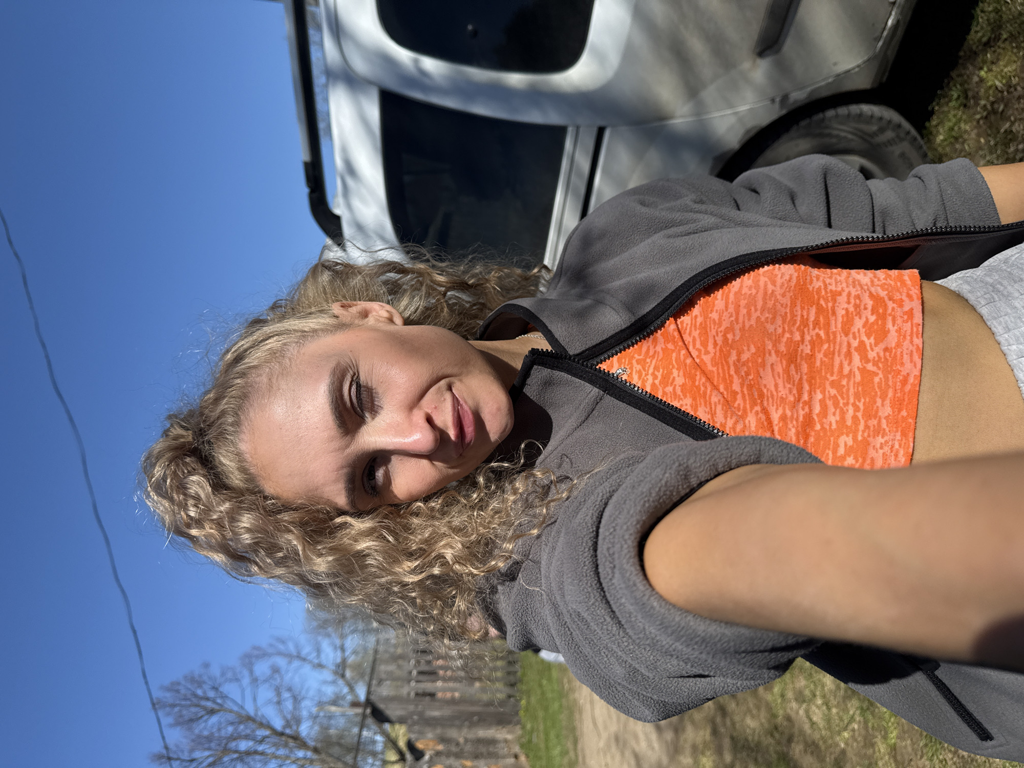 A blonde woman is posing for a selfie outdoors in Nizhnyaya Chermoda, Russia. She has curly blonde hair and is wearing a gray zip-up fleece jacket over an orange patterned top. A white vehicle is visible in the background, with a tire partly in the frame. The sky is blue and there are trees visible in the distance. A weathered wooden fence is also noticeable. The ground has patches of dry grass and dirt.