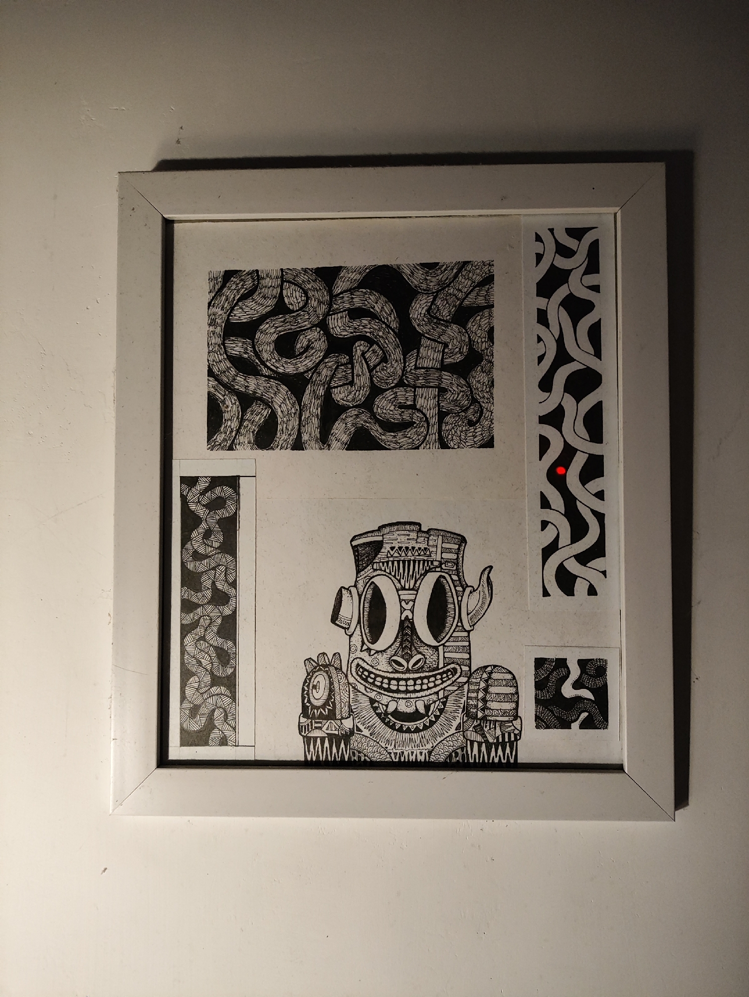 A framed black and white ink drawing is displayed on a white wall. The drawing features a central cartoonish figure with large eyes, sharp teeth, and horns, surrounded by intricate patterns of wavy lines. The artwork is divided into several sections, with abstract patterns in the top and left sections, and smaller patterned squares on the right. A single red dot is visible on the right-hand side of the drawing, possibly a reflection or part of the artwork. The wall appears to be textured and lit from above, casting shadows on the right side of the frame. The location is described as Kepung, Indonesia, however, there are no visual elements in the image to confirm or deny this specific location.