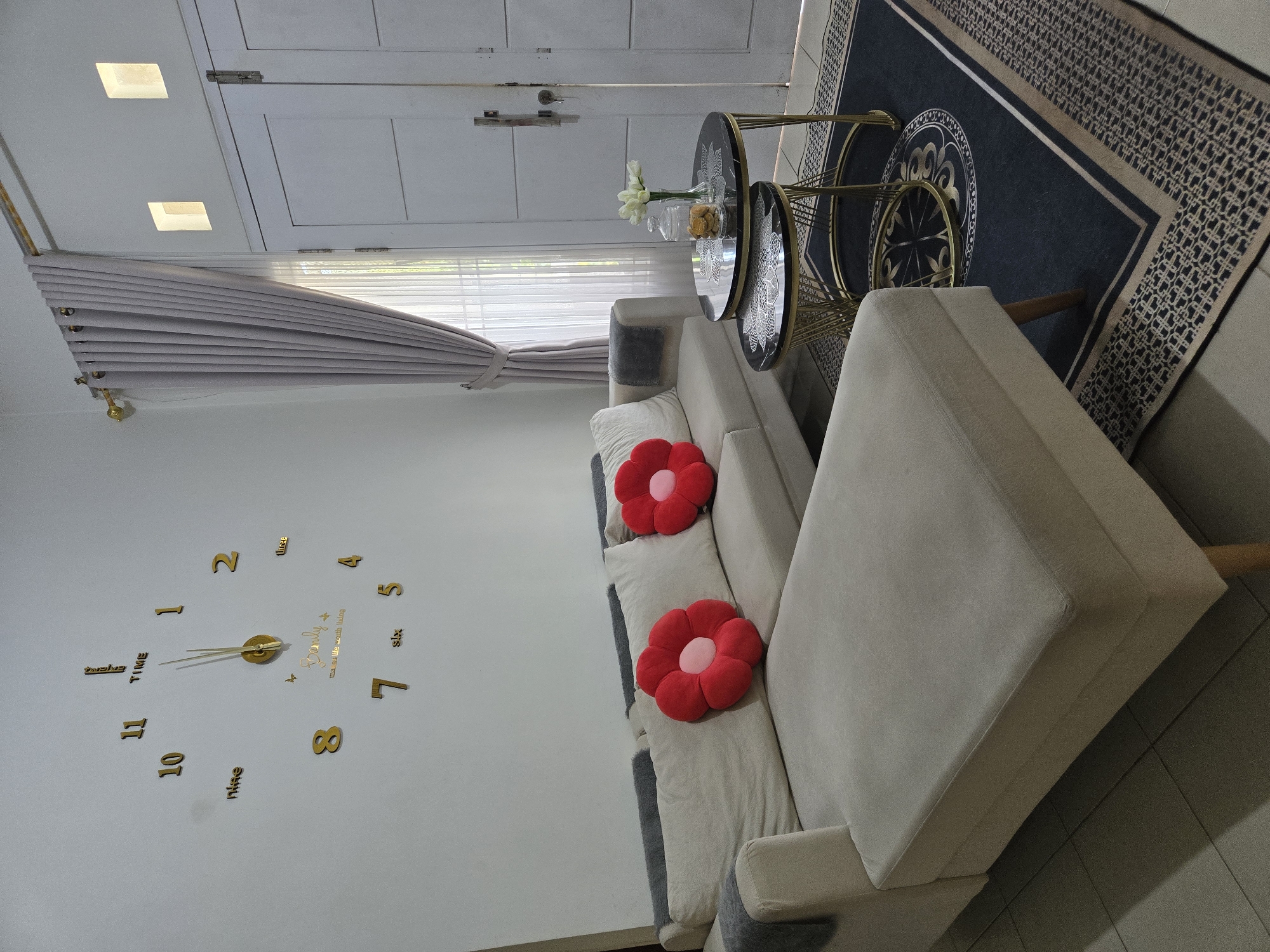 The image captures an indoor scene, likely a living room, with a modern aesthetic. Dominating the scene is a large, cream-colored sectional sofa with two red flower-shaped throw pillows. To the left, a large decorative wall clock with gold numerals and a quote, "Family makes life worth living," is prominently displayed. In the center right, a set of nesting coffee tables with black marble tops and gold wire bases is arranged on a dark rug with a patterned border. A glass jar filled with cookies and a vase with white flowers are on the top table. Behind the coffee tables, a tall white door with two panels is visible, flanked by a light grey curtain that partially covers a window. Two small, rectangular light fixtures are set into the wall above the door. The flooring appears to be tiled. The overall impression is of a clean, well-decorated, and comfortable living space. This scene is situated in Sungailiat, Indonesia.