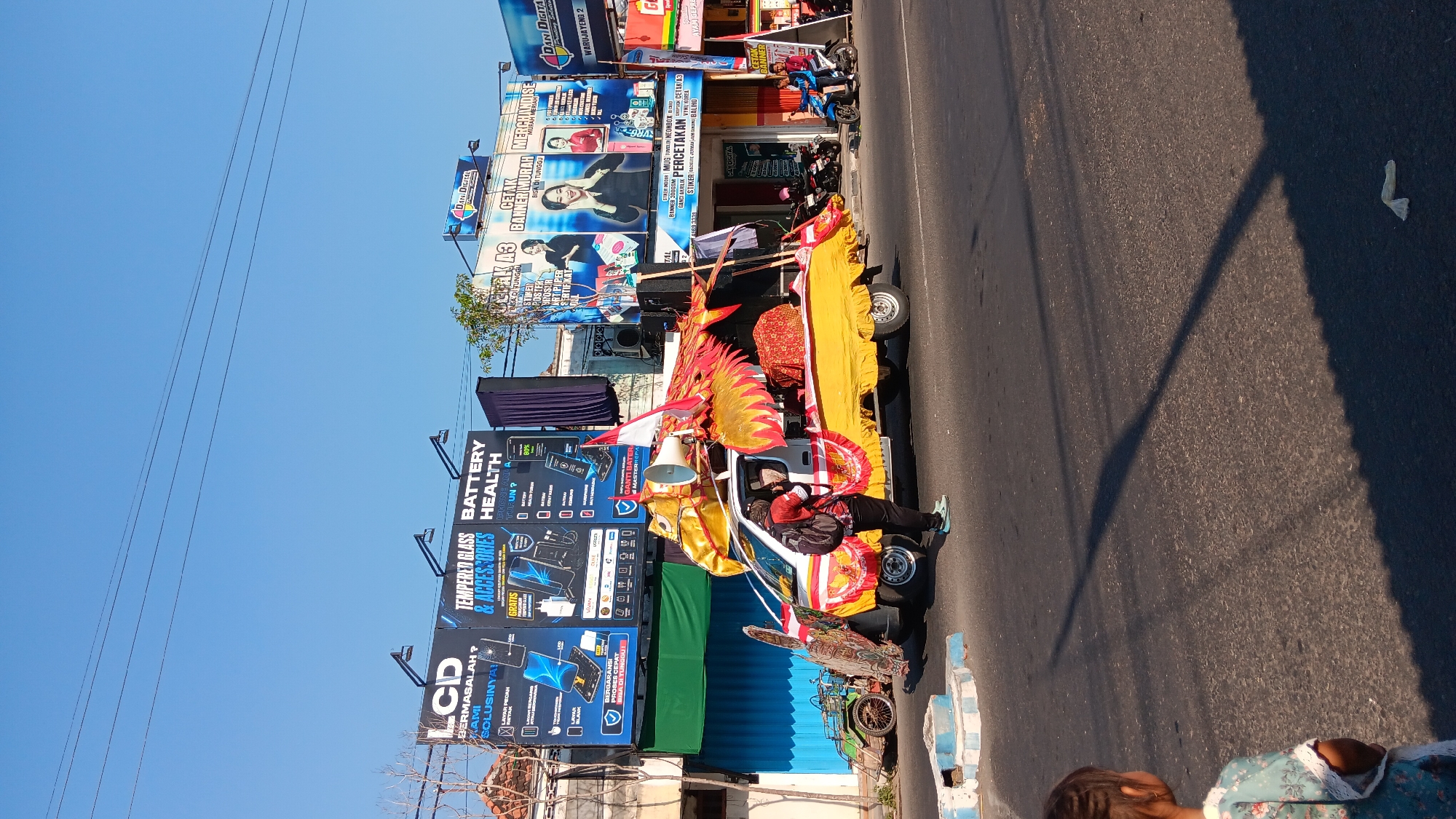 A person stands on the street next to a brightly decorated yellow pick-up truck that appears to be part of a parade or celebration. The truck is adorned with a large, golden dragon-like structure with red accents, and a large speaker is mounted on its roof. A small green vehicle is parked on the left side of the road. In the background, several billboards are visible, advertising various services and products, including LCDs, tempered glass, battery health, and printing services. The billboards are in Indonesian. The scene is set on a sunny day with a clear blue sky. The location is identified as Warujayeng, Indonesia, based on one of the visible billboards. A young child is visible in the lower left corner of the frame, looking away from the camera. Several motorcycles are parked in front of a building in the background.
