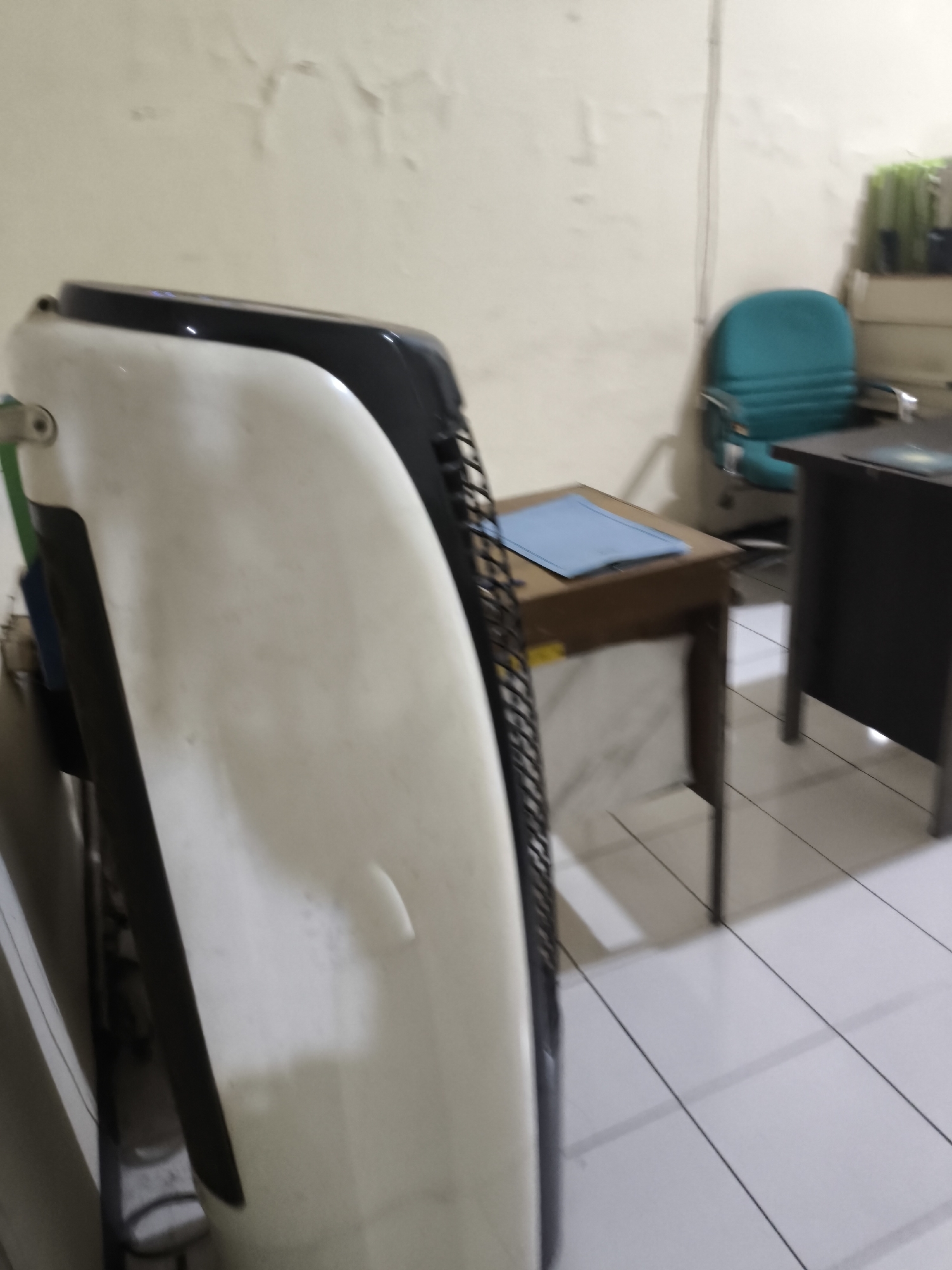 The image shows an indoor scene, likely an office or meeting room, in Cilegon, Indonesia. A large, white and black object, possibly an air conditioner or a piece of furniture, dominates the foreground and is angled towards the viewer. In the background, there are two desks, one dark brown wooden desk with a blue mat on it, and a darker desk behind it. A teal office chair is positioned near the wooden desk. The walls are a light, pale color with some peeling paint. The floor is tiled in a light color. On a shelf in the far background, there are some green plants in containers. The lighting appears to be artificial, casting shadows on the floor.