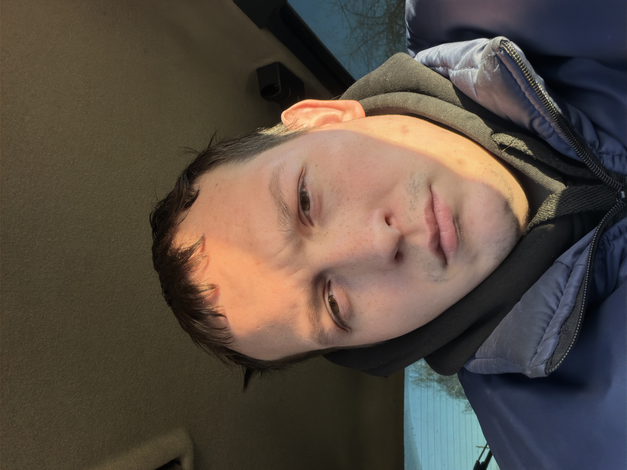 A man is taking a selfie. He is wearing a black hoodie and a blue jacket. The interior of a vehicle is visible in the background. Outside the car, a building and tree branches are visible. The photo was potentially taken in the city of Chaikovsky, Russia.