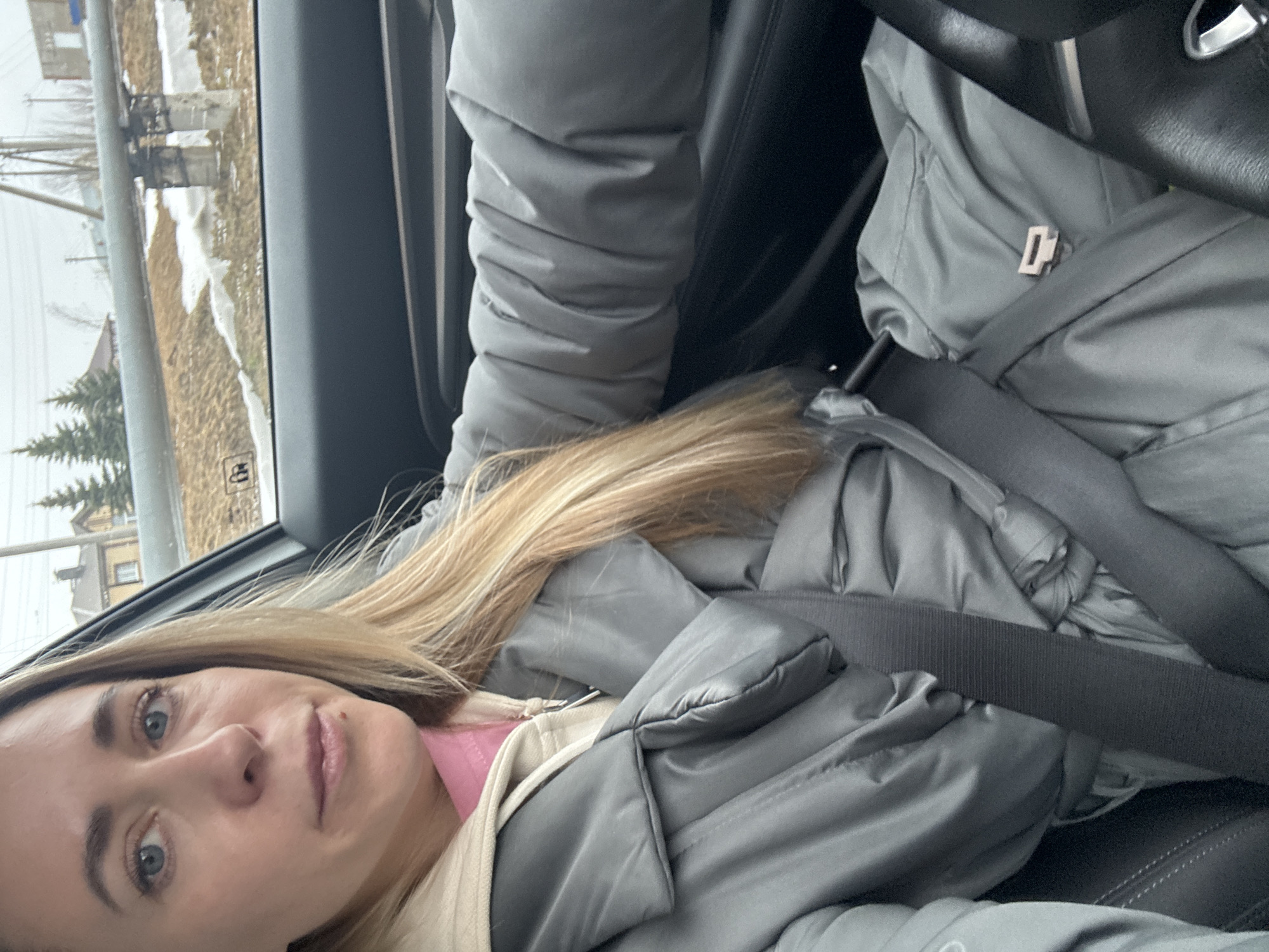 A woman is wearing a grey puffy jacket and a seatbelt in the driver's seat of a car. The car is moving. Through the window, a view of Chaikovsky, Russia is visible, including a snow-covered landscape, trees, and buildings.
