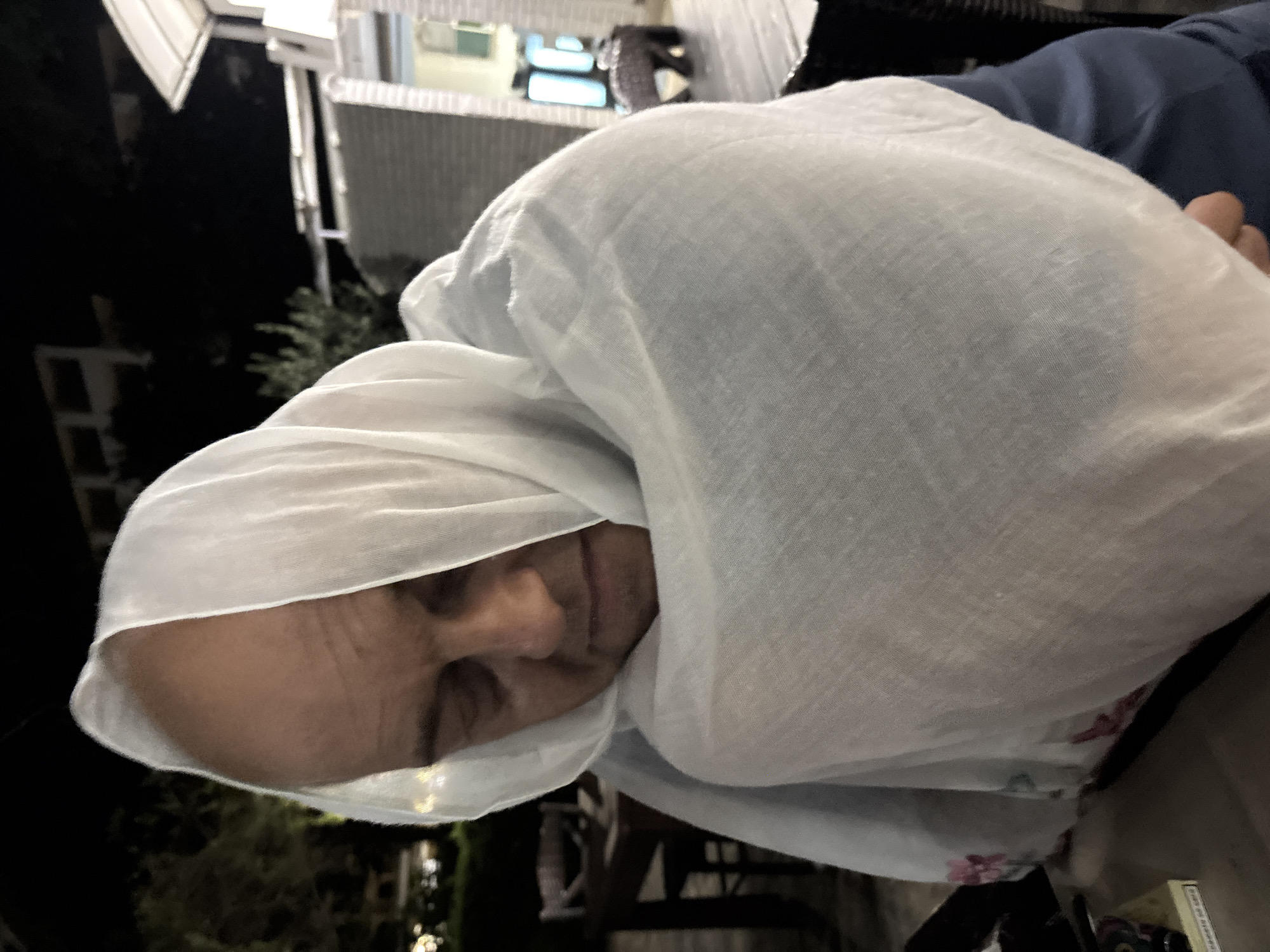 A bald man is wrapped in a white shawl outdoors at night, with his eyes closed and head bowed. The scene appears to be in a restaurant or outdoor seating area, with tables and chairs visible in the background, along with some greenery and building structures. The man is wearing a dark blue shirt under the shawl. The lighting suggests artificial light sources. The location is identified as Varna, Bulgaria.