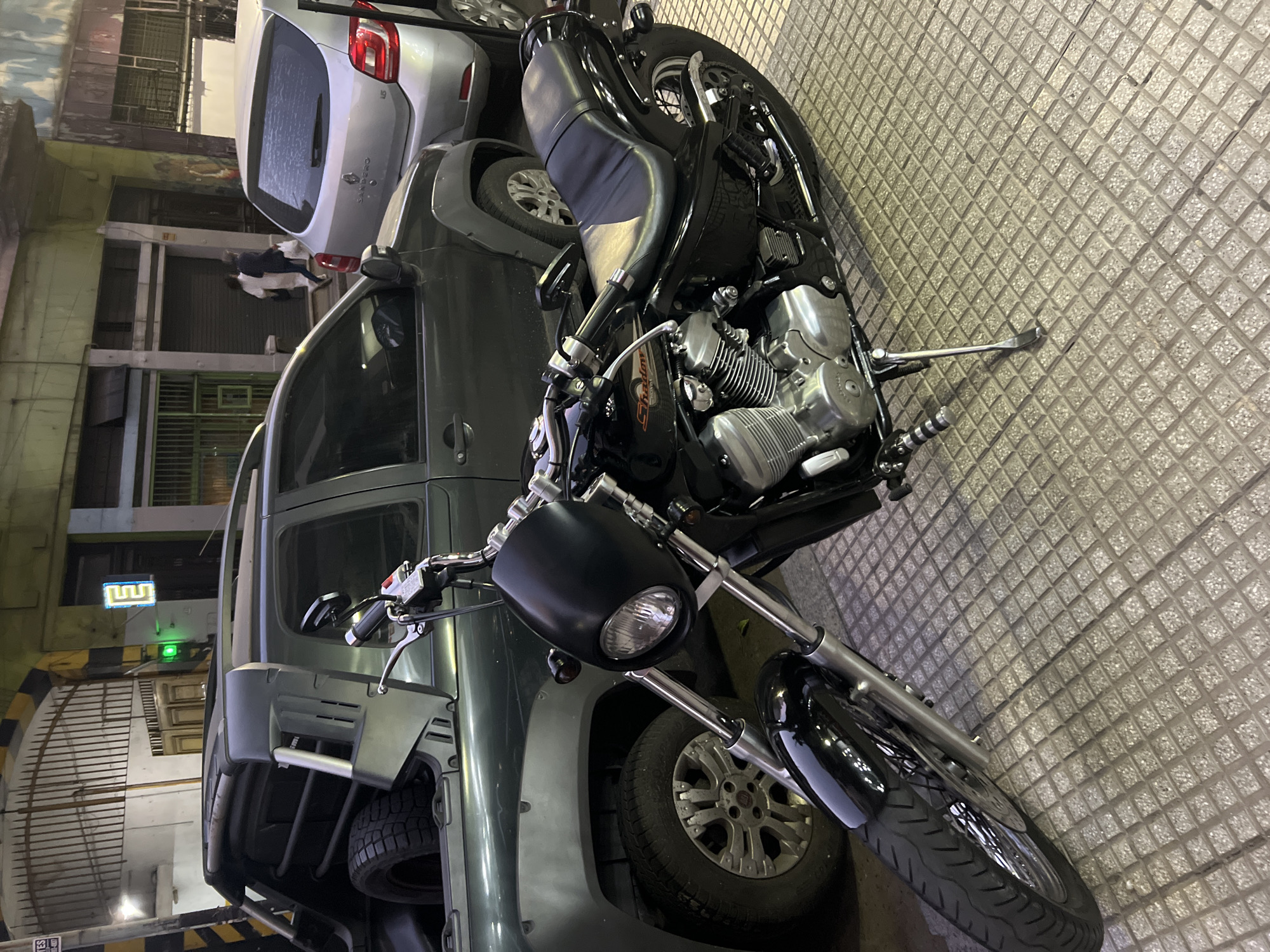 An evening scene captures a black cruiser-style motorcycle parked on a textured, square-patterned paved surface in Buenos Aires, Argentina. The motorcycle, identifiable by the "Shadow" text on its tank, features a matte black headlight housing, chrome handlebars, and an engine with prominent cooling fins. Its black leather seat and overall design suggest a classic custom aesthetic.
Behind the motorcycle, a dark grey SUV or pickup truck, possibly a Fiat Toro, is parked, with its rear section and spare tire visible. Further back, a silver Renault Sandero hatchback is parked, clearly showing "Sandero" and the Renault logo on its rear. The vehicles are positioned in what appears to be a parking area or a street-side spot next to buildings.
In the background, dimly lit buildings rise, indicating night or late evening. A small figure, possibly a person, is visible between two buildings in the distance. A distinct green illuminated sign, shaped like a stylized 'M' or Greek key pattern, adds a touch of light to the upper left background. On the paved ground in the foreground, near the bottom left of the overall image, a universal "No Parking" sign (a red circle with a diagonal line) is faintly visible. The scene is static, depicting parked vehicles and urban architecture.