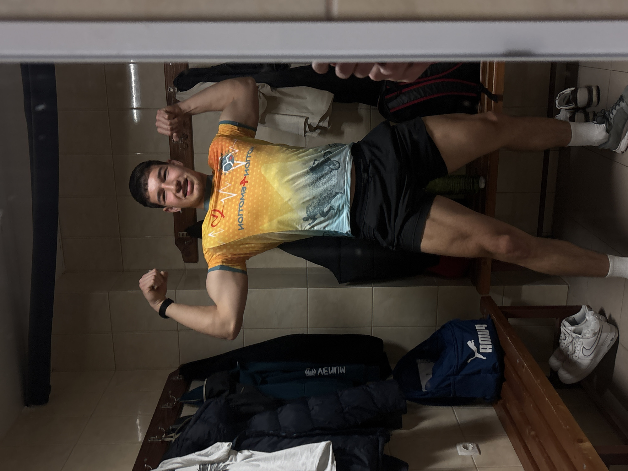 The image shows a man posing in a mirror. He is wearing a short-sleeved yellow and blue shirt and black shorts. He is flexing both arms. He is wearing a black fitness tracker on his left wrist. He has short dark hair. The reflection shows he is in a room with a wooden bench and a coat rack. Clothes, bags and shoes are on the bench and under it. The floor is tiled. The man's location in Zlatograd, Bulgaria cannot be determined from the given image.