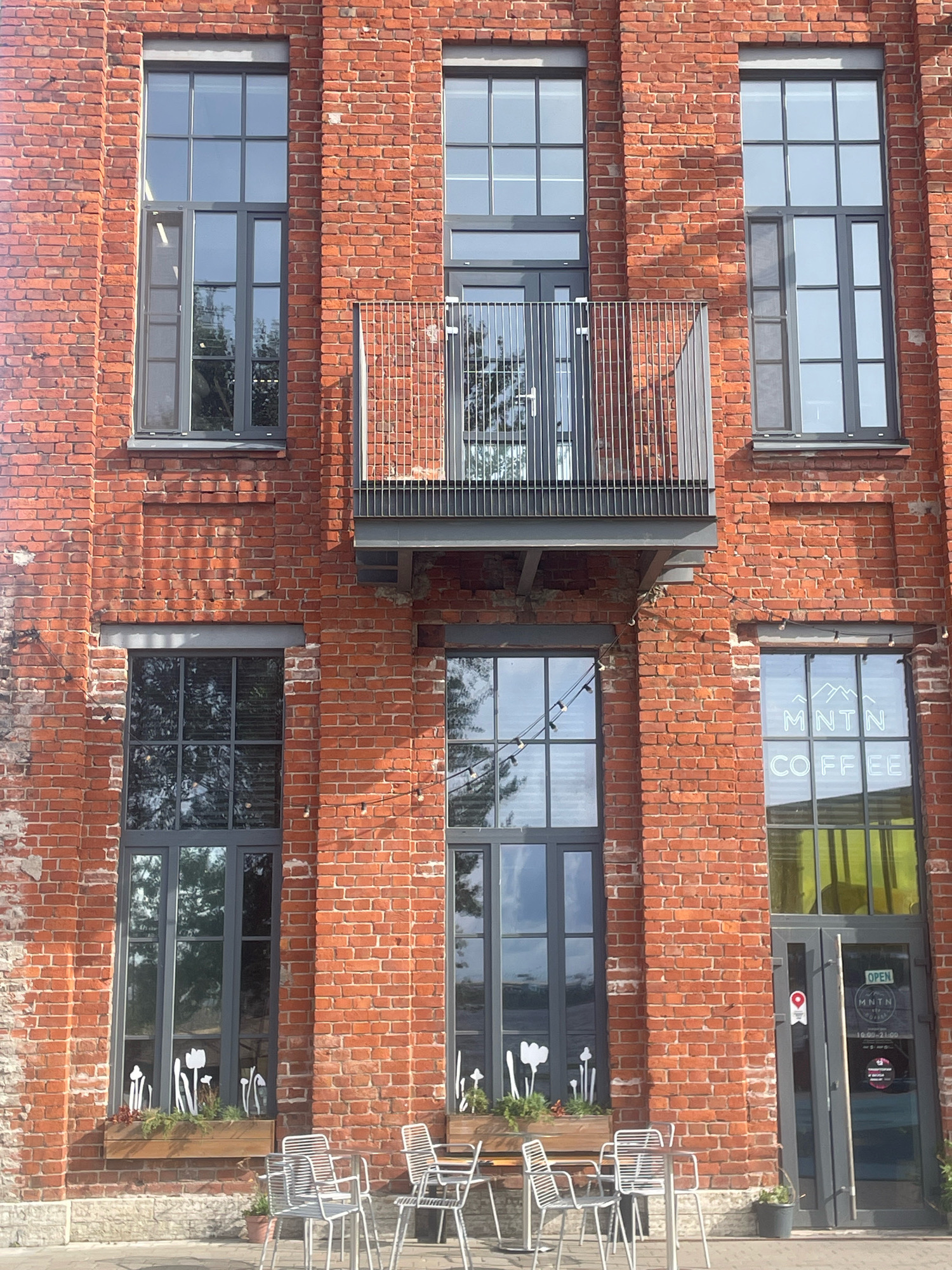 A close-up, eye-level, outdoor medium shot captures the brick facade of a building in Saint Petersburg, Russia. The building features multiple tall, dark-framed windows with a grid pattern, and a metal balcony with vertical railings is visible on the upper level. String lights are suspended across some of the windows. Below, a planter box with white tulip decorations sits in front of one window, and several empty metal chairs and tables are arranged on the pavement. A glass door with the "OPEN" sign and "MNTN COFFEE" logo is visible on the right side of the image. The brickwork shows signs of age and weathering. The overall scene suggests the exterior of a cafe or commercial establishment.