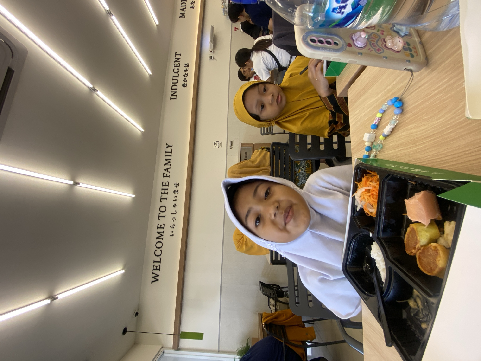 In a brightly lit indoor setting, likely a restaurant in Sidoarjo, Indonesia, two children wearing headscarves (one white and one yellow) are seated at a wooden table. A bento box containing various foods (rice, shredded carrots, and what appear to be grilled meat and rolled omelet) is visible in front of the child in the white headscarf. An adult's hand is visible near the yellow headscarf. A water bottle is on the table near a cellphone with charms attached. The wall bears the inscriptions "WELCOME TO THE FAMILY," "INDULGENT," and "MADE," along with corresponding text in Japanese. Other people are seated in the background. The scene suggests a family outing to a restaurant in Sidoarjo, Indonesia.