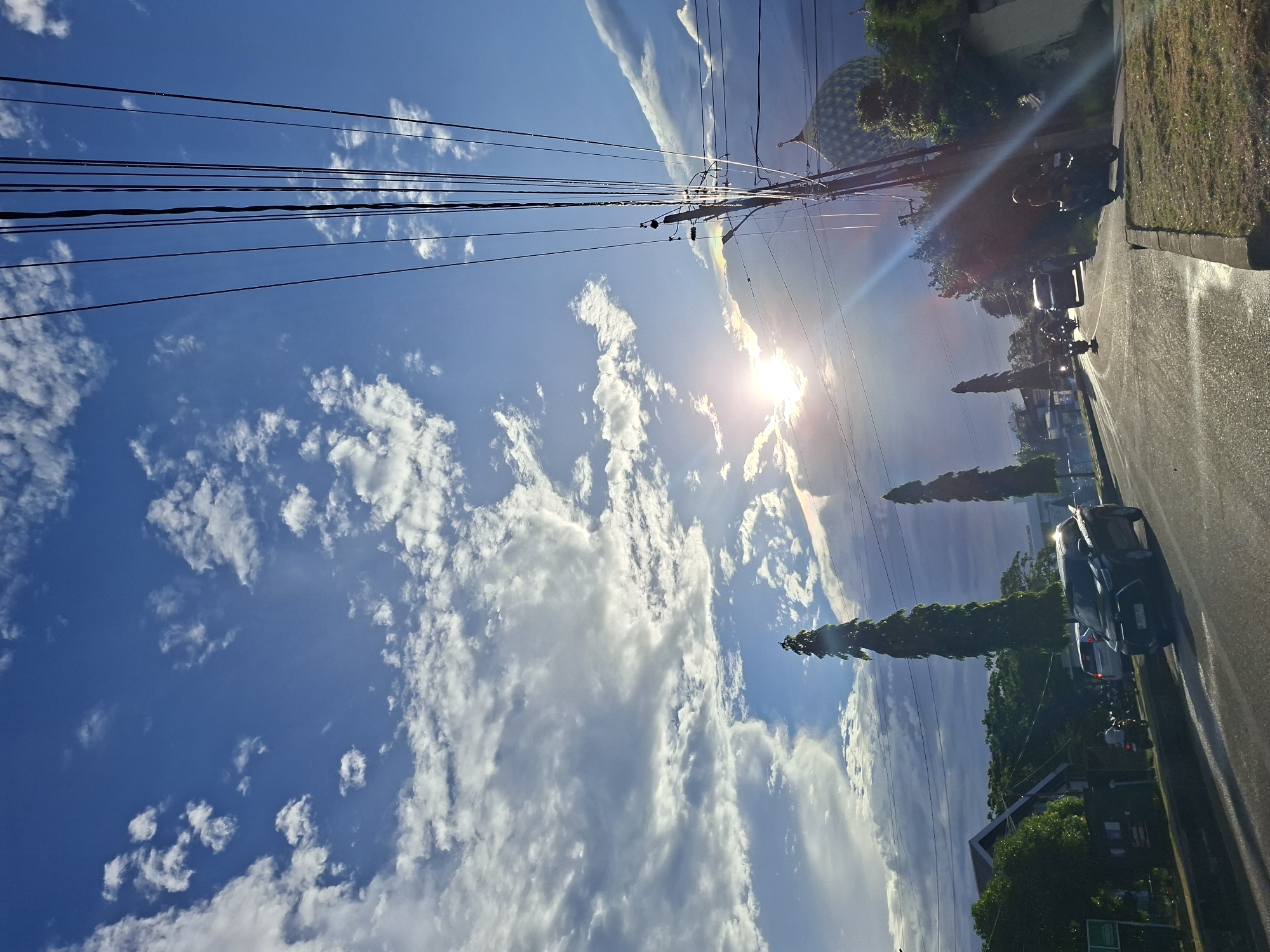 The image is a street-level shot in Lhokseumawe, Indonesia, under a bright blue sky filled with cumulus clouds. The sun shines intensely, creating lens flare. Power lines stretch across the upper portion of the frame, supported by a utility pole. A street runs along the right side, with parked cars and motorcycles. Tall, narrow trees line the street. A building with a patterned dome-like structure is partially visible in the upper right.
