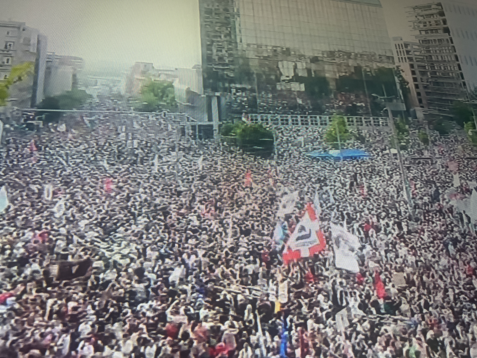 A large crowd of people fills a public space in what appears to be a city center. Buildings surround the area, including modern high-rises with reflective glass facades and older, low-rise structures. Many people in the crowd are holding flags and signs.
Based on the visible features, this image seems to depict a gathering in Sofia, Bulgaria. The buildings in the background match the architectural styles found in Sofia's city center. Further investigation of specific landmarks would confirm the exact location within the city.
