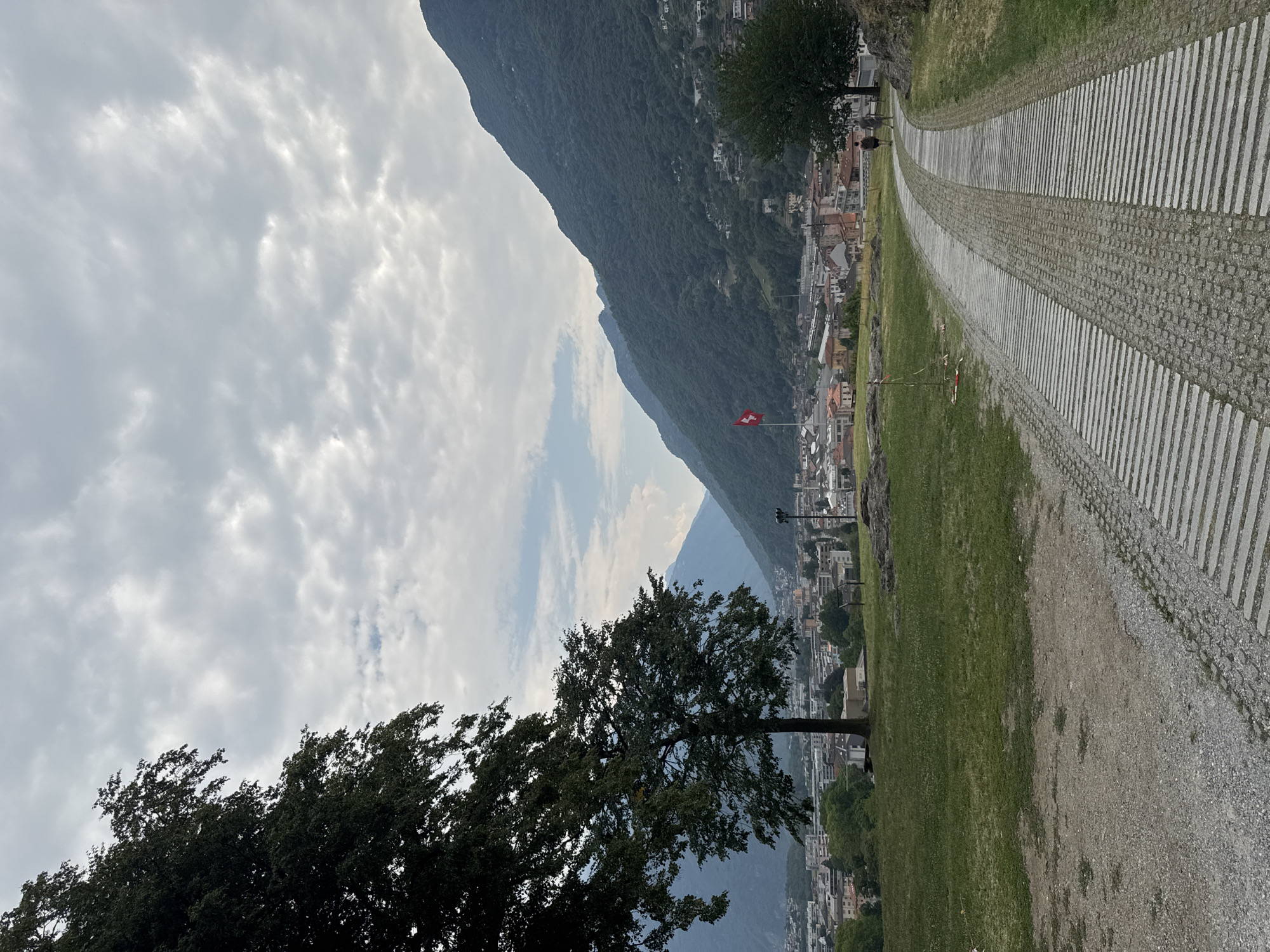 This is a wide-angle, eye-level shot of a pathway in Bellinzona, Switzerland, looking out over the city and surrounding mountains. The pathway in the foreground is paved with a mixture of gravel and large, rectangular stones laid horizontally. Two people are walking away from the camera on this pathway. To the left of the pathway is a grassy slope dotted with rocks. In the middle ground, a mature tree with dark green foliage stands prominently on the grassy area, with the city of Bellinzona spread out behind it. The city consists of multi-story buildings with red-tiled roofs. A Swiss flag is visible on a flagpole in the distance, set against the backdrop of steep, tree-covered mountains. The sky is overcast with dramatic clouds. The scene appears to be a public park or viewpoint overlooking the city.