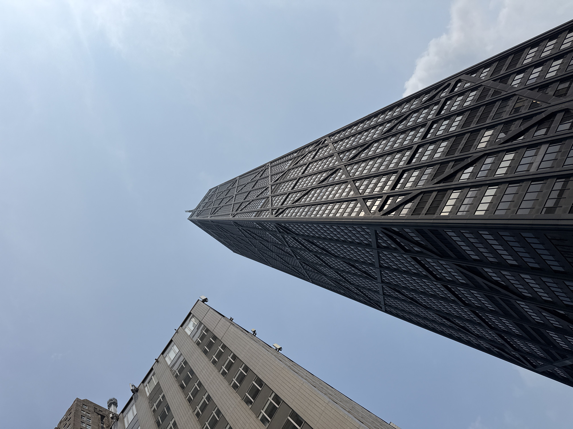 The image shows a low-angle, wide shot of two skyscrapers against a clear, pale blue sky with some wispy clouds. On the left is a tall, grey building with a grid of windows. On the right is a much taller, dark skyscraper with a distinctive exoskeleton structure and a multitude of windows. The two buildings are angled towards each other, creating a dramatic perspective. This is a view of the John Hancock Center (now known as 875 North Michigan Avenue) in Chicago, Illinois, United States of America.