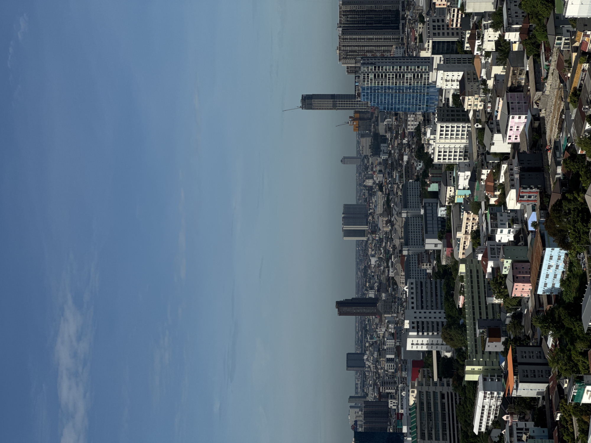 This is a high-angle, full shot of the cityscape of Bangkok, Thailand. The image showcases a dense urban environment with a mix of residential and commercial buildings, some of which are high-rise. The sky above is a pale blue with scattered wispy clouds. In the mid-ground, several tall, modern buildings, including skyscrapers, are visible. One skyscraper under construction features a crane. In the foreground, lower-rise buildings and a significant amount of greenery, likely trees, dominate the landscape, suggesting a well-established residential area. The lighting indicates it is daytime, possibly late morning or early afternoon, with the sun casting distinct shadows. There are no people or discernible actions visible in the image.