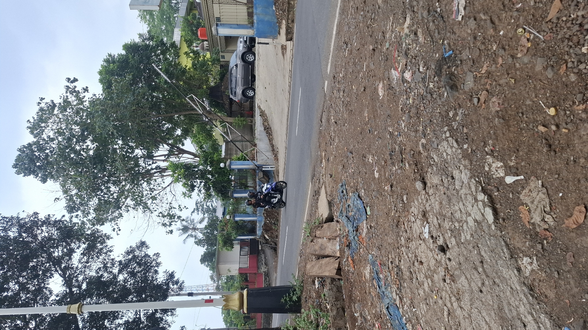 The image depicts a roadside scene in Cilaku, Indonesia. In the foreground, the ground is covered in dirt, rocks, and scattered debris, including pieces of blue plastic sheeting and dry leaves. A damaged section of asphalt is visible. A dark pole with a golden finial stands on the left side of the image.
In the mid-ground, a paved road runs horizontally. Two people are riding a blue and white motorcycle on the road, heading away from the viewer. The driver and passenger are wearing dark clothing and helmets.
Beyond the road, a blue and white fence borders a property. A grey SUV is parked on the right side of the property, near a yellow building. There are trees and some structures with red roofs in the background. The sky is overcast.