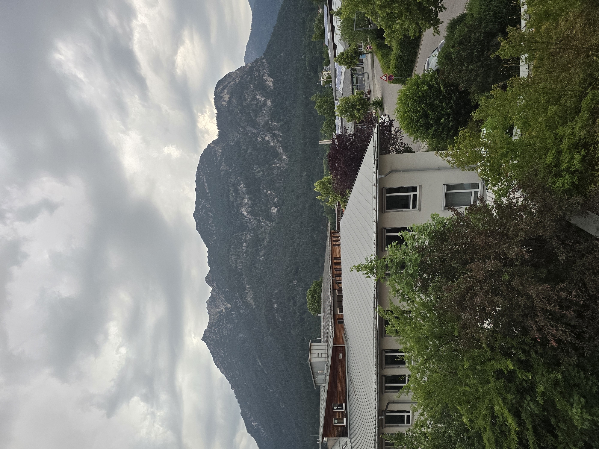 The image depicts a view of the town of Garmisch-Partenkirchen, Germany, dominated by a large, heavily wooded mountain range under a cloudy sky. In the foreground, a modern-looking building with a white facade and gray metal roof is visible, featuring several windows and some greenery in front of it. Adjacent to this building are other structures, some with wooden accents and more gray metal roofs. The middle ground shows a road with a parked white car and a yield sign. The background is filled with the imposing mountain, its slopes covered in dense forest. The overall atmosphere is overcast and subdued.