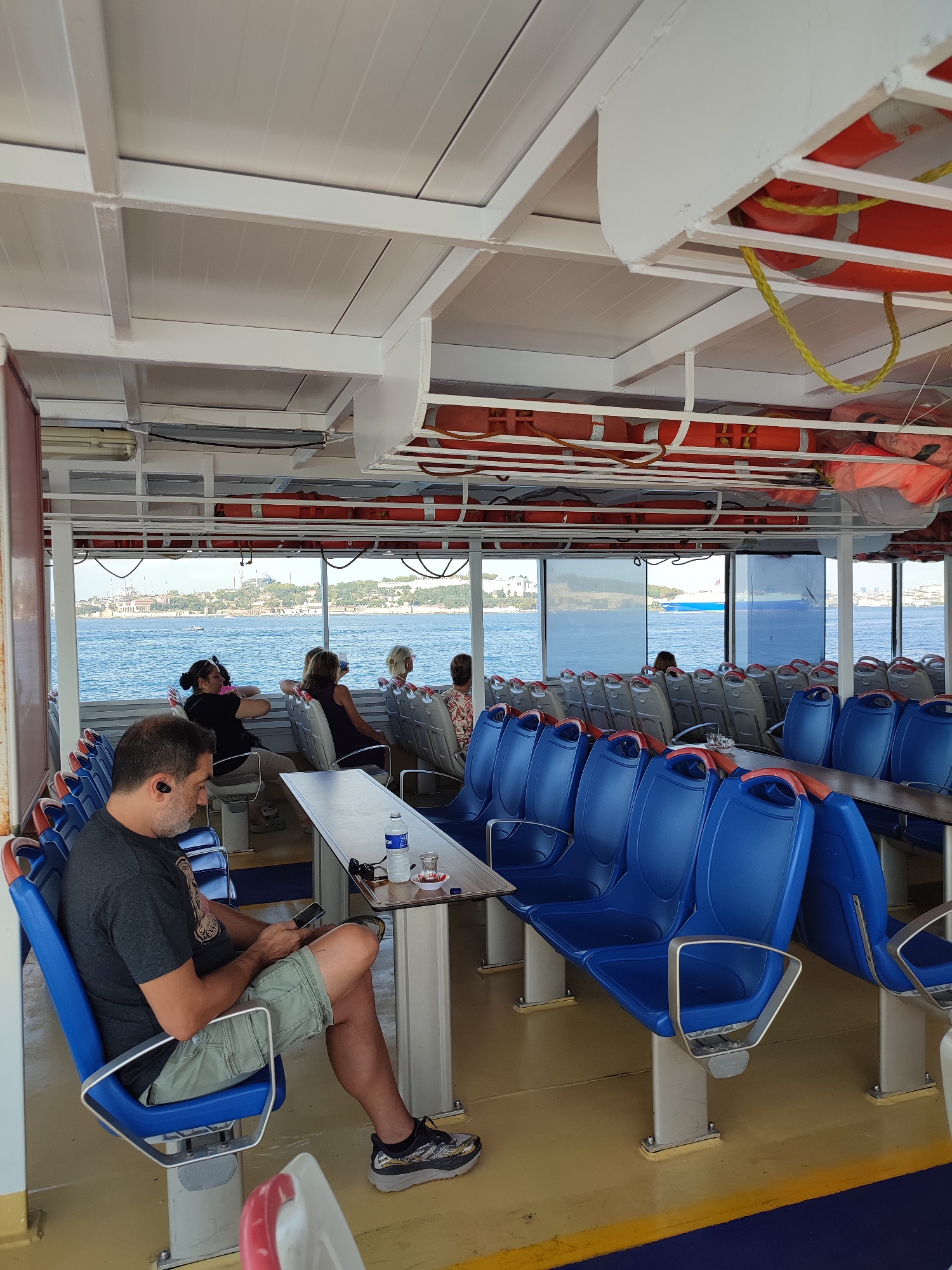 An indoor view from a ferry traveling on the Bosphorus strait near Üsküdar, Turkey, shows several passengers seated on blue and gray seats. A man in the foreground, wearing a dark t-shirt and khaki shorts, is looking at his phone. He has black earbuds in his ears. A table in front of him holds a bottle of water and a small glass of tea. In the background, through the windows, the Istanbul skyline is visible, including the Hagia Sophia and the Blue Mosque. Other passengers are seated further down the ferry, facing the windows. Orange life preservers are stored overhead. The ferry's interior features white panels and metal beams.
