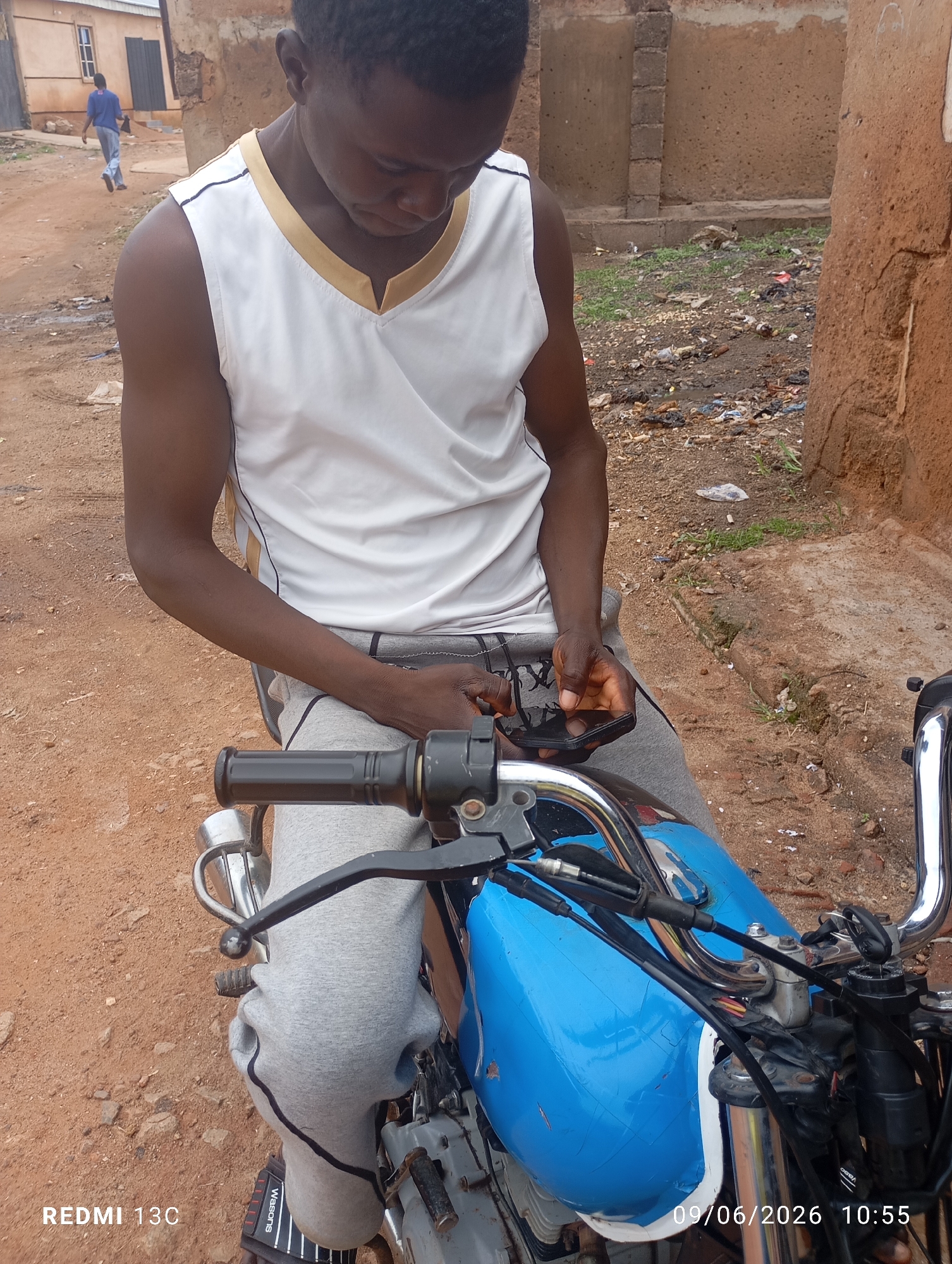 A young Black male wearing a white sleeveless shirt with gold trim and grey sweatpants is sitting on a blue motorcycle. He is looking down at a black smartphone in his hands. The motorcycle is parked on a dirt path in front of a dilapidated building. In the background, another person is walking away from the camera. The environment appears to be an urban area with scattered debris. The date and time stamp on the image reads "09/06/2026 10:55".