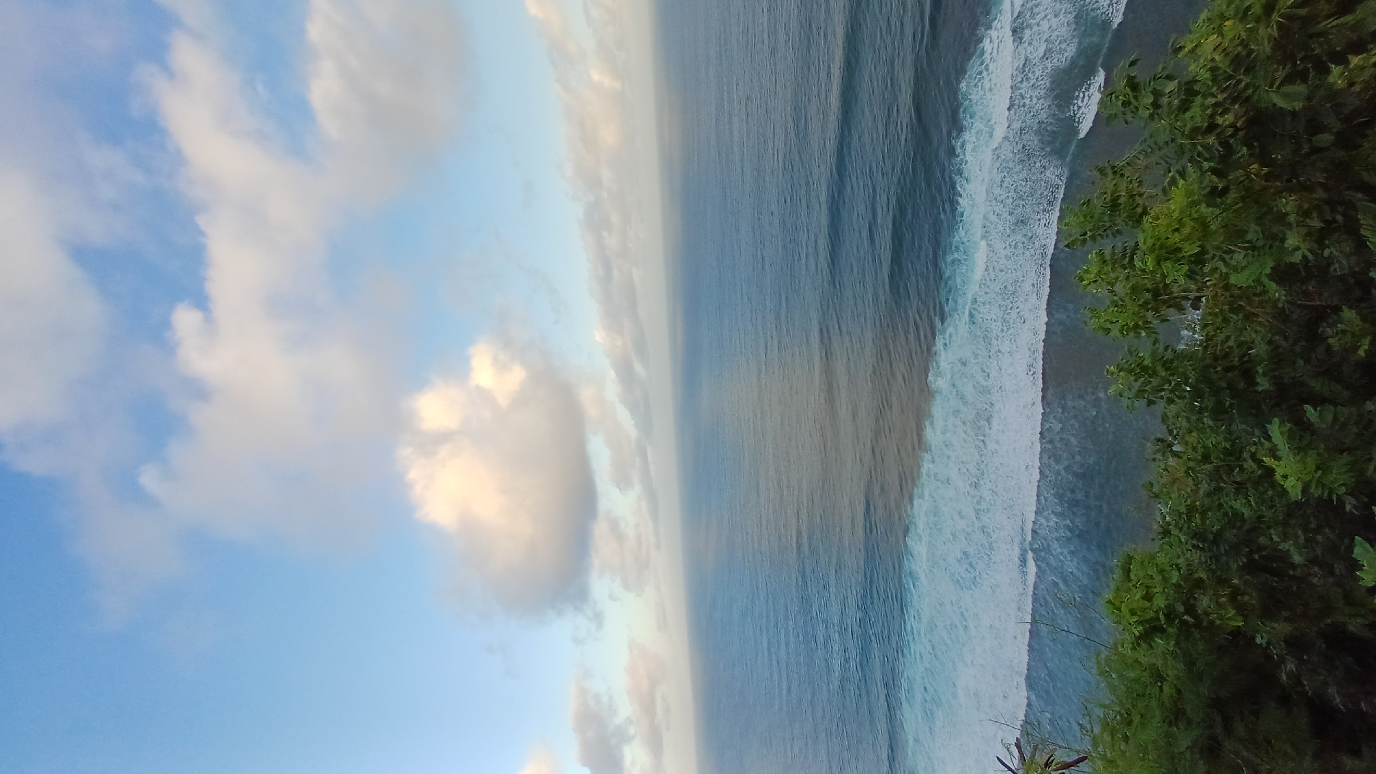 The image shows a view from a cliff overlooking the ocean in Kuta Selatan, Indonesia. The sky is blue with scattered clouds, which are illuminated by the setting sun. Below, the dark blue ocean stretches out to the horizon, with waves crashing against the base of the cliff. Green foliage covers the edge of the cliff.