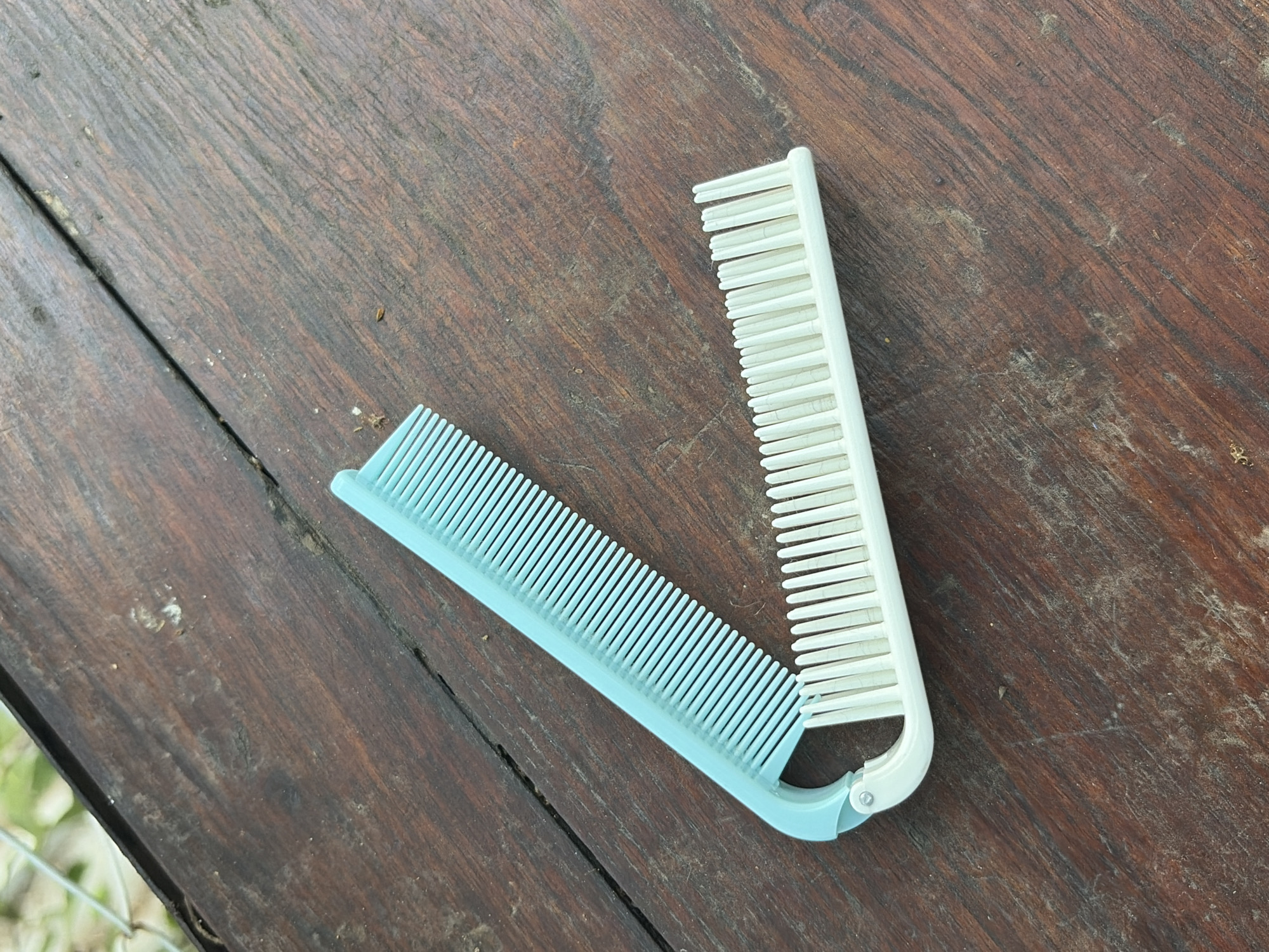 A pair of light blue and white plastic combs are laid open on a wooden surface. The light blue comb is positioned in the upper left of the frame, angled downwards and to the right. The white comb is positioned below and to the right of the blue comb, angled upwards and to the right, with the two combs forming a 'V' shape. The wooden surface appears to be a table or deck, with visible grain patterns and some debris. The background is blurred and shows greenery, possibly foliage. The scene is outdoors, and based on the context provided, the location is the city of Pekalongan, Indonesia.