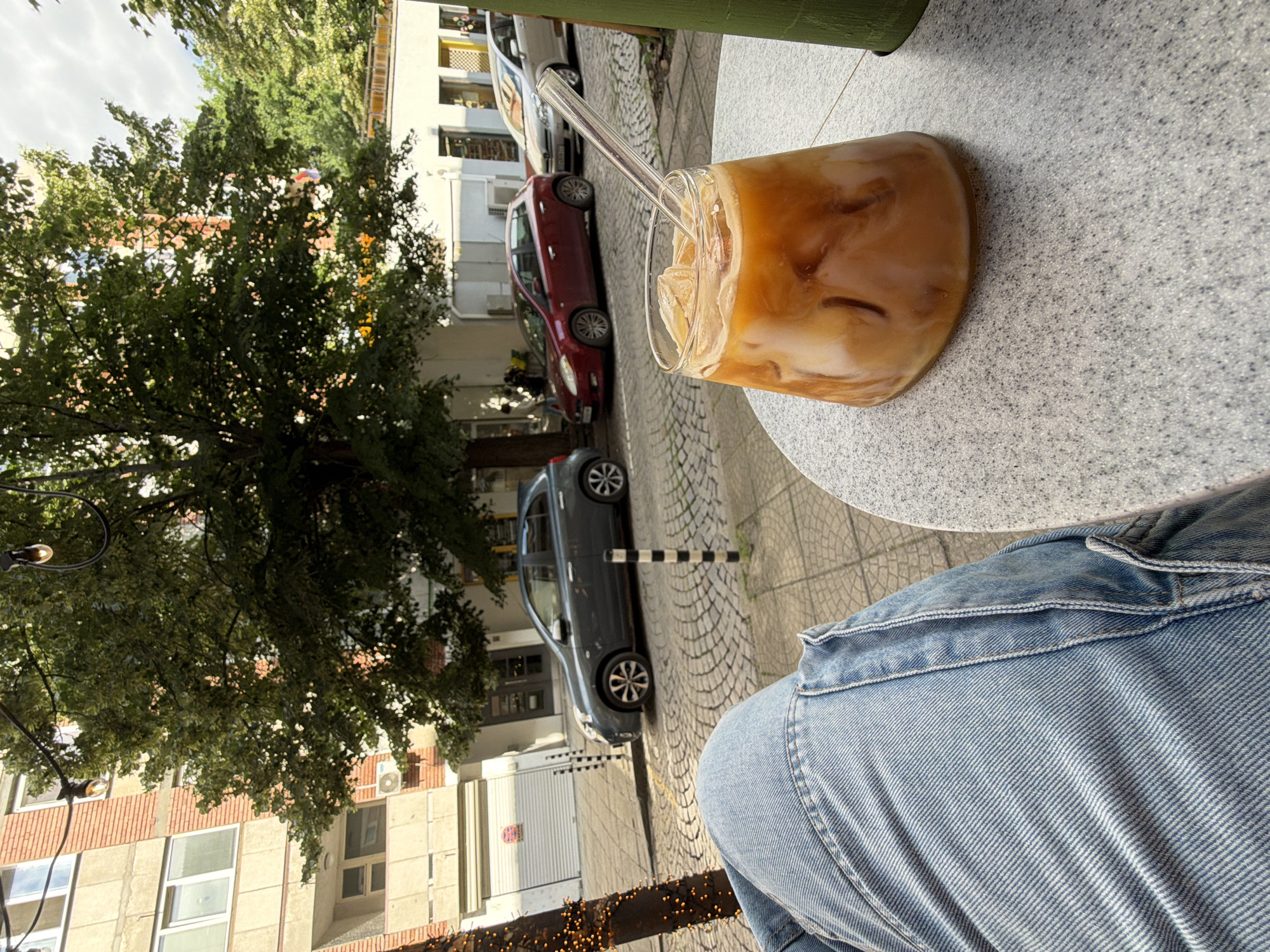 The media file is a photograph taken from a person's perspective, looking down and to the side. The foreground is dominated by the person's legs, clad in light blue denim jeans. A glass of iced coffee with milk is prominently featured on a speckled grey table, with a clear glass straw emerging from it. The coffee is a light brown color with visible ice cubes and swirls of milk.
In the background, a tree with lush green foliage partially obscures a street scene. Several cars are parked along a cobblestone street. One car is a dark grey sedan, another is a red hatchback, and further back, a white car is visible. Buildings with multiple windows and brick accents line the street. The overall impression is that of a casual outdoor setting, likely a cafe or restaurant patio, during the daytime, as suggested by the bright lighting and shadows. The weather appears to be pleasant, possibly sunny. The image conveys a relaxed and leisurely atmosphere. There is no visible text in the image.