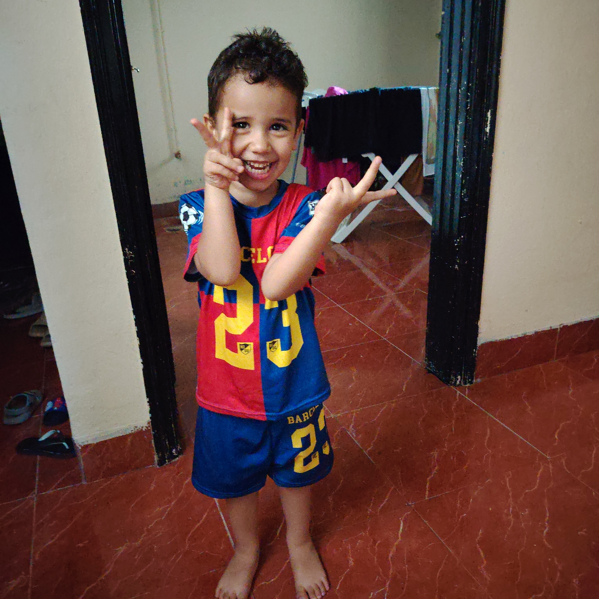 A young boy, appearing to be around 5-7 years old, stands in a doorway, smiling widely and making a "peace sign" gesture with both hands. He is wearing a red and blue soccer jersey with the number "23" in yellow on the front and blue shorts with the word "BARC" and the number "23" also in yellow. The boy's feet are bare, and he is standing on a reddish-brown tiled floor. The background shows a hallway with a black-framed doorway on the left and a tan wall on the right. In the background, through an open doorway, a clothes drying rack with laundry is visible. The scene is set indoors, likely in a residential area of Ouaouizert, Morocco, indicated by the architectural style and flooring commonly found in Moroccan homes. The lighting suggests an indoor environment, possibly illuminated by natural light from an adjacent room or artificial lighting not directly visible in the frame.