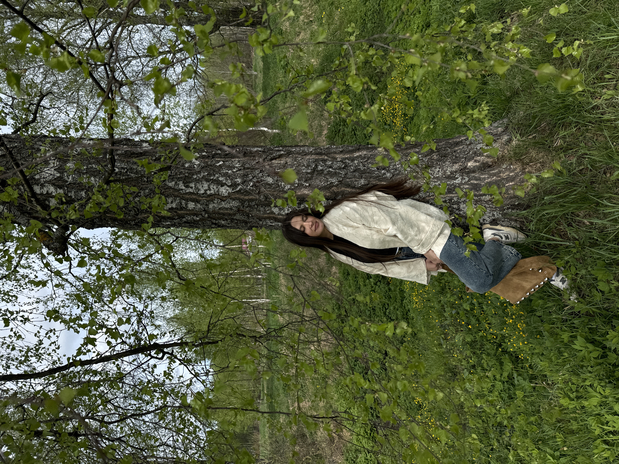 A young woman is lying on the trunk of a birch tree in a grassy area in Красный Маяк, Russia. The tree trunk runs horizontally across the middle of the image. The woman is wearing a light-colored jacket and blue jeans and is lying with her head towards the left side of the frame. Green foliage surrounds the tree trunk and extends into the background. The sky is visible through the branches of the tree.