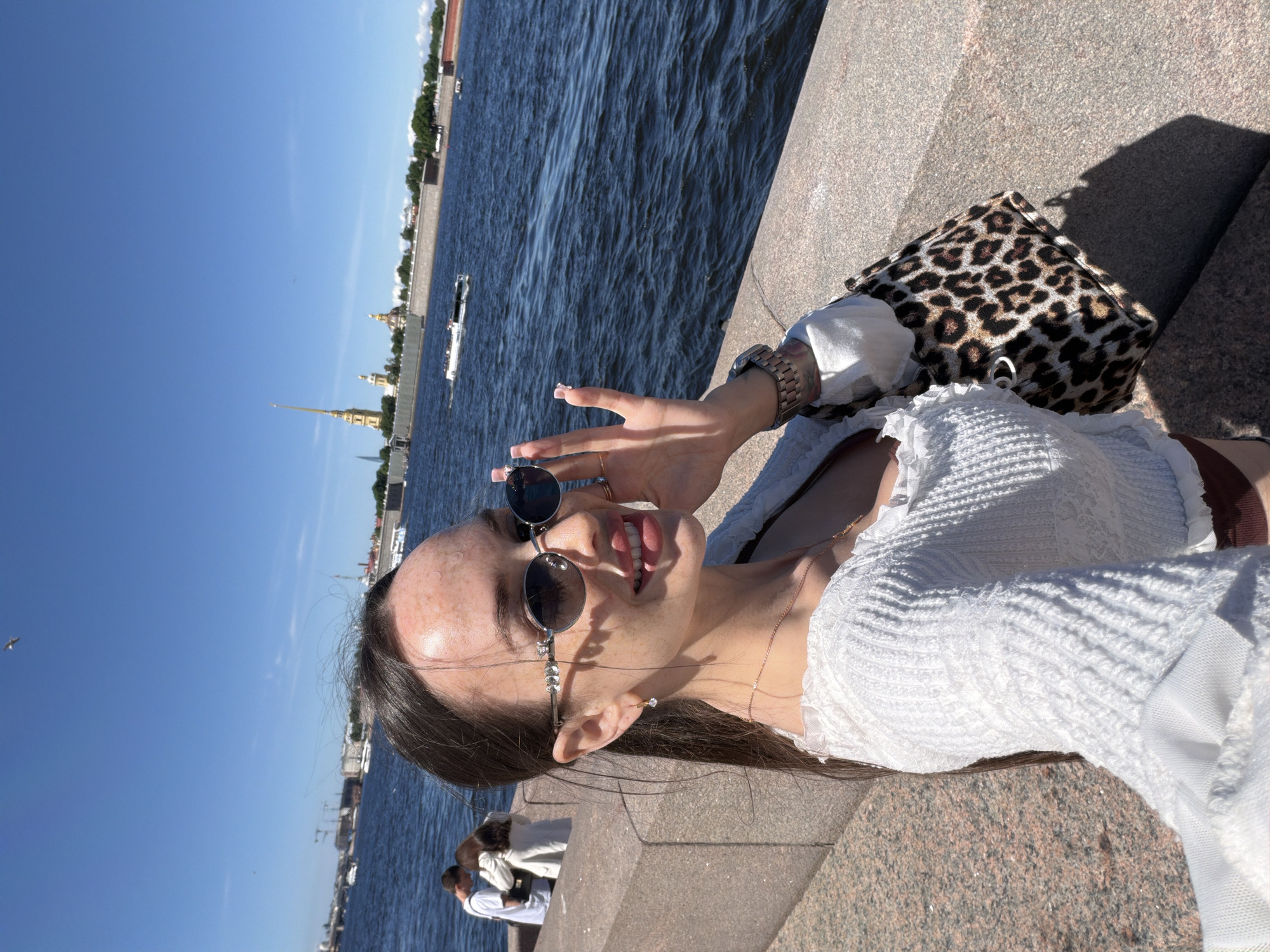 The image depicts a young woman taking a selfie on the granite embankment of the Neva River in Saint Petersburg, Russia, on a clear, sunny day. The **main subject**, a woman with long, dark hair pulled back, is positioned in the lower right and center of the frame, looking towards the camera with a smile. She has fair skin with numerous freckles across her forehead, nose, and cheeks. She wears oval-shaped sunglasses adorned with clear crystals on the frames, and her left hand is raised to her face, partially obscuring the sunglasses and her cheek, with manicured nails visible. She is dressed in a white, textured, long-sleeved top with ruffled details. A thin gold chain necklace and a small, clear-stone earring are visible on her left side. A metallic-band watch is on her left wrist, and a leopard-print tote bag hangs on her right side, partially visible.
In the **background** to the left, two individuals stand near the embankment, facing away from the camera; one wears a white t-shirt, and the other a white jacket. The **environment** features a vibrant blue sky with minimal cloud cover. The wide body of water, the Neva River, occupies the middle ground, showing small ripples and at least two white passenger boats moving in the distance. The **cityscape** across the river includes the iconic golden spire of the Peter and Paul Cathedral and other domed structures on Vasilyevsky Island or the Peter and Paul Fortress. Various buildings, some with cranes, line the distant riverbanks, interspersed with greenery. The foreground is dominated by the reddish-brown granite blocks of the embankment, which extends along the river. Sunlight from the top right casts distinct shadows, including that of the woman's bag, onto the embankment.
