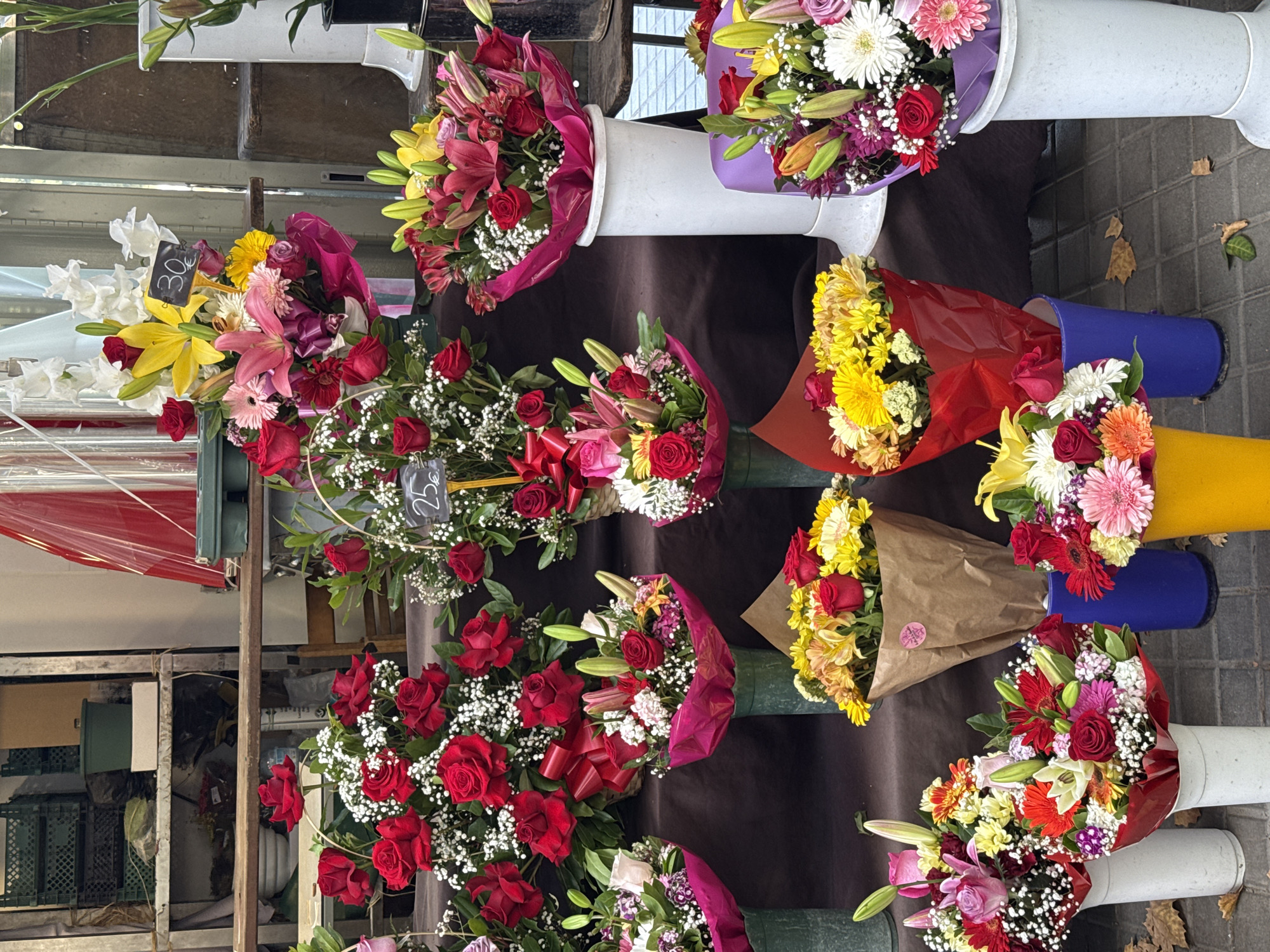 The image depicts a vibrant flower stall, likely a street vendor, showcasing an abundant display of various floral arrangements. The dominant colors are reds, pinks, yellows, and whites, reflecting the diversity of the flowers. Several bouquets are presented in vases or wrapped in decorative paper. Some arrangements include roses, lilies, and chrysanthemums, interspersed with smaller white flowers and greenery. Price tags are visible on some of the bouquets, with one reading "30€". The stall appears to be set up outdoors, with a tiled ground surface visible and what seems to be part of a building or structure in the background. The overall impression is one of a well-stocked and visually appealing flower market. Without specific architectural or landmark clues, pinpointing the exact location within Barcelona is not possible from the visual information alone.