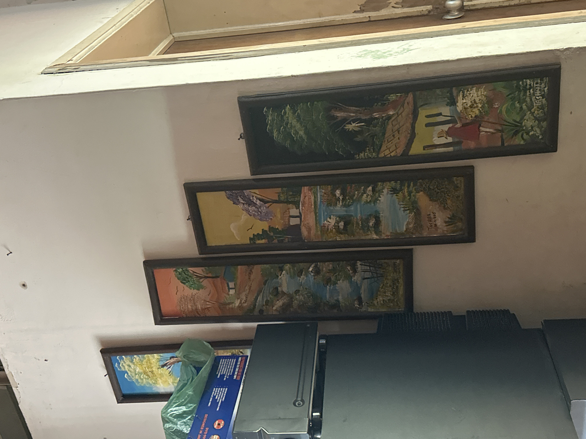 The image depicts an indoor scene in Juan Griego, Venezuela, featuring a light-colored wall adorned with five framed landscape paintings, a stack of electronic equipment, and a partially open wooden doorway.
On the left, a dark-framed painting of a yellow-leafed tree against a blue sky is partially obscured by a crumpled green plastic bag resting on a dark grey electronic device. This device, possibly a television or monitor, has a visible side speaker grille and a circular button/indicator on its front, and is stacked upon another dark grey electronic unit. A label on the top unit is partially visible, reading "DESIGNED IN JA" and "DVD-999-DA".
To the right of the electronic equipment, four vertically oriented, dark-framed landscape paintings are arranged along the wall. The first, from the left, shows a house, trees, and a waterfall under an orange sky. The second depicts a similar scene with a house, trees, and a waterfall under a yellow sky, with a visible signature "TACHIRA 10-11-2006" at the bottom. The third painting, partially visible, features a large green tree against a dark background, with a partial signature "TACHIRA". The fourth and final painting on the far right depicts a figure in a red garment and hat holding a stick, walking past a house with a thatched roof and flowering plants, also with a visible signature "TACHIRA" in the lower right corner.
The light-colored wall exhibits minor imperfections and small hooks. To the far right, a light-colored door frame encloses a partially open wooden door, revealing a glimpse of an adjacent room and a light-colored doorknob. The ceiling is visible in the upper left corner.