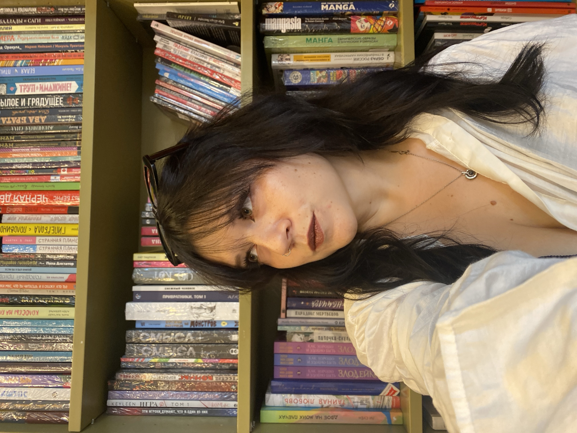 A young woman with dark, wavy hair and brown eyes is photographed from the chest up, looking to her right. She is wearing a white button-down shirt that is unbuttoned to reveal her chest and a silver necklace with a circular pendant. Her hair is partially obscuring her face. She has a nose ring and dark lipstick. Her glasses are perched on top of her head.
The woman is standing in front of a bookshelf filled with books. The books appear to be various genres and sizes, with some spines showing Cyrillic lettering, suggesting a location in Russia. The overall setting appears to be a library or a personal study. The lighting is natural and comes from the front, illuminating the woman's face and the books. The background is slightly blurred, placing emphasis on the woman. There are no other people or animals visible in the image. The colors in the image are muted, with the white of the woman's shirt and the varied colors of the book spines being the most prominent.