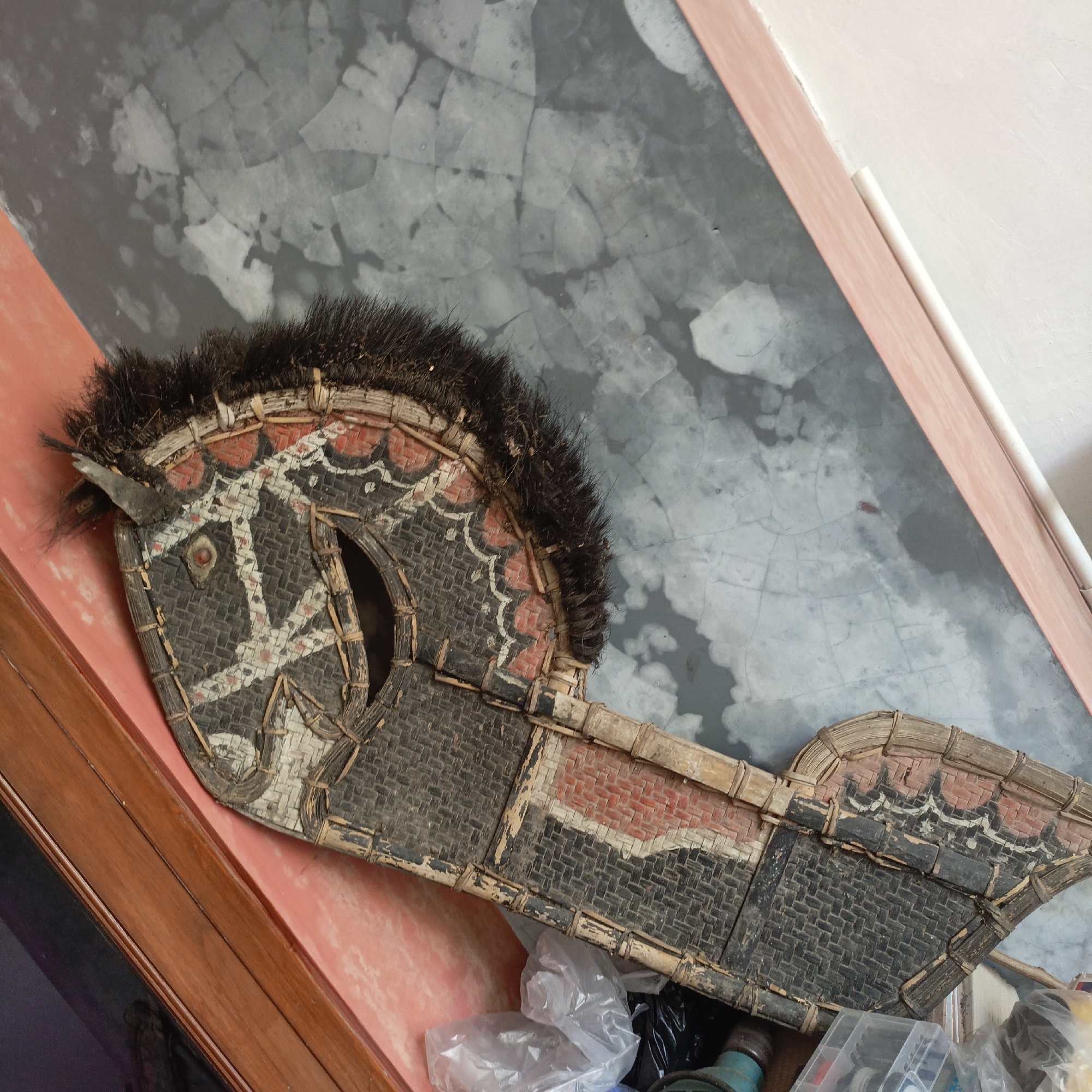 A close-up, angled shot shows a traditional Indonesian horse puppet, likely a "Kuda Lumping" or "Jathilan" prop, resting on a surface. The puppet, constructed from woven bamboo and adorned with black, red, and white patterns, features a mane of dark, coarse hair. Its eye is a small red bead. The background is a textured gray wall with visible cracks, partially covered by a pinkish border and a white pipe. To the lower right, plastic bags and containers are visible, suggesting a storage area. The puppet appears to be made of natural materials and has a rustic, aged appearance. It is likely located in Salatiga, Indonesia, possibly in a home or workshop where such crafts are made or stored.