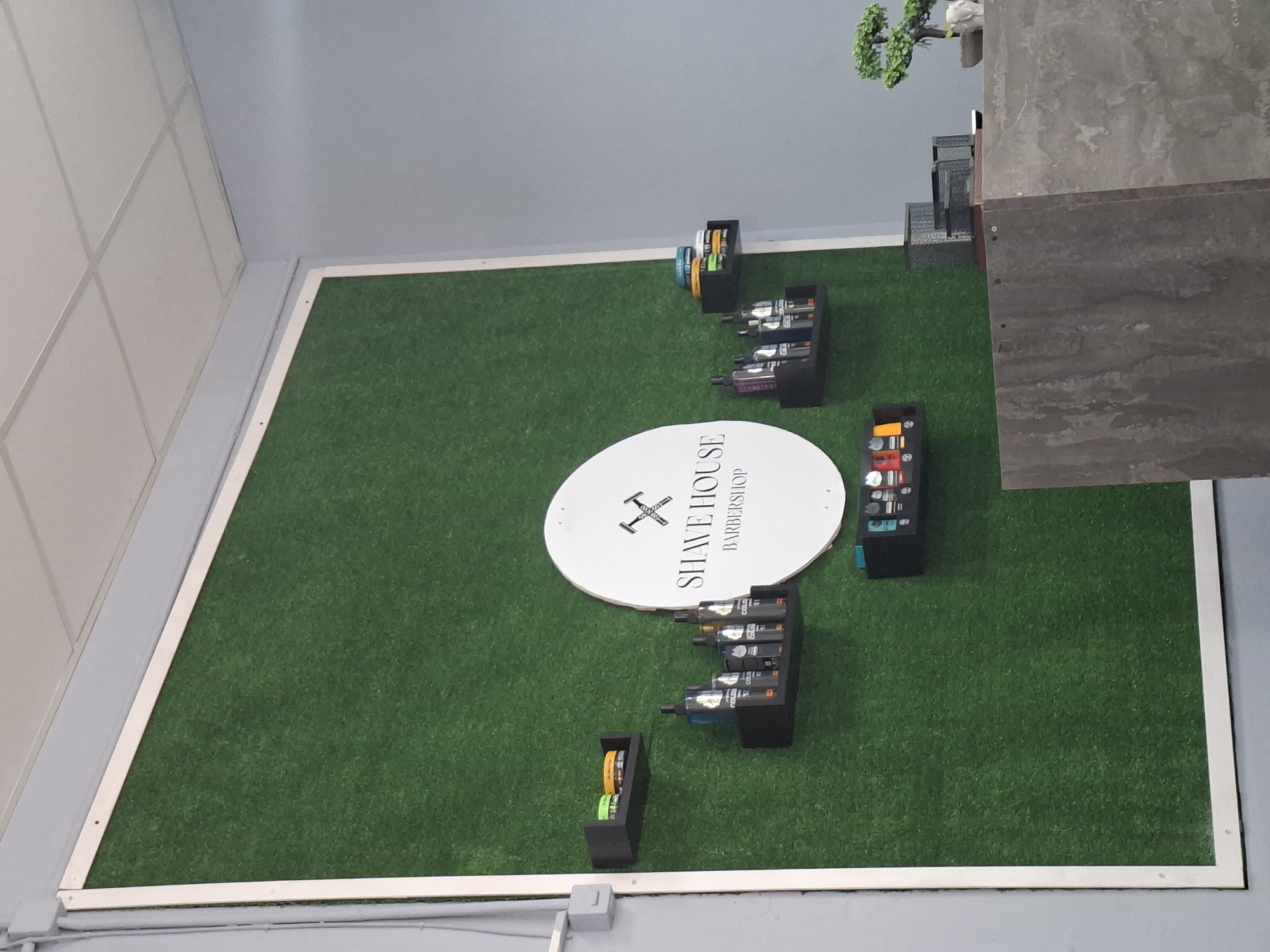 A slightly overhead, tilted shot depicts an indoor area, likely a business or retail space, located in Galabovo, Bulgaria. The floor is covered in artificial green turf, bordered by a white-trimmed, light grey baseboard.
The center of the frame features a white circular sign that reads "SHAVE HOUSE BARBERSHOP" with an abstract icon above the text. Various black display stands holding product containers are arranged on the turf.
Along the upper edge of the frame, against a light blue-grey wall, is a small, decorative tree with white accents. Adjacent to it is a tiered grey object, potentially a display structure. The room is well-lit, though the source isn't visible. A white tiled ceiling is on the left of the shot.