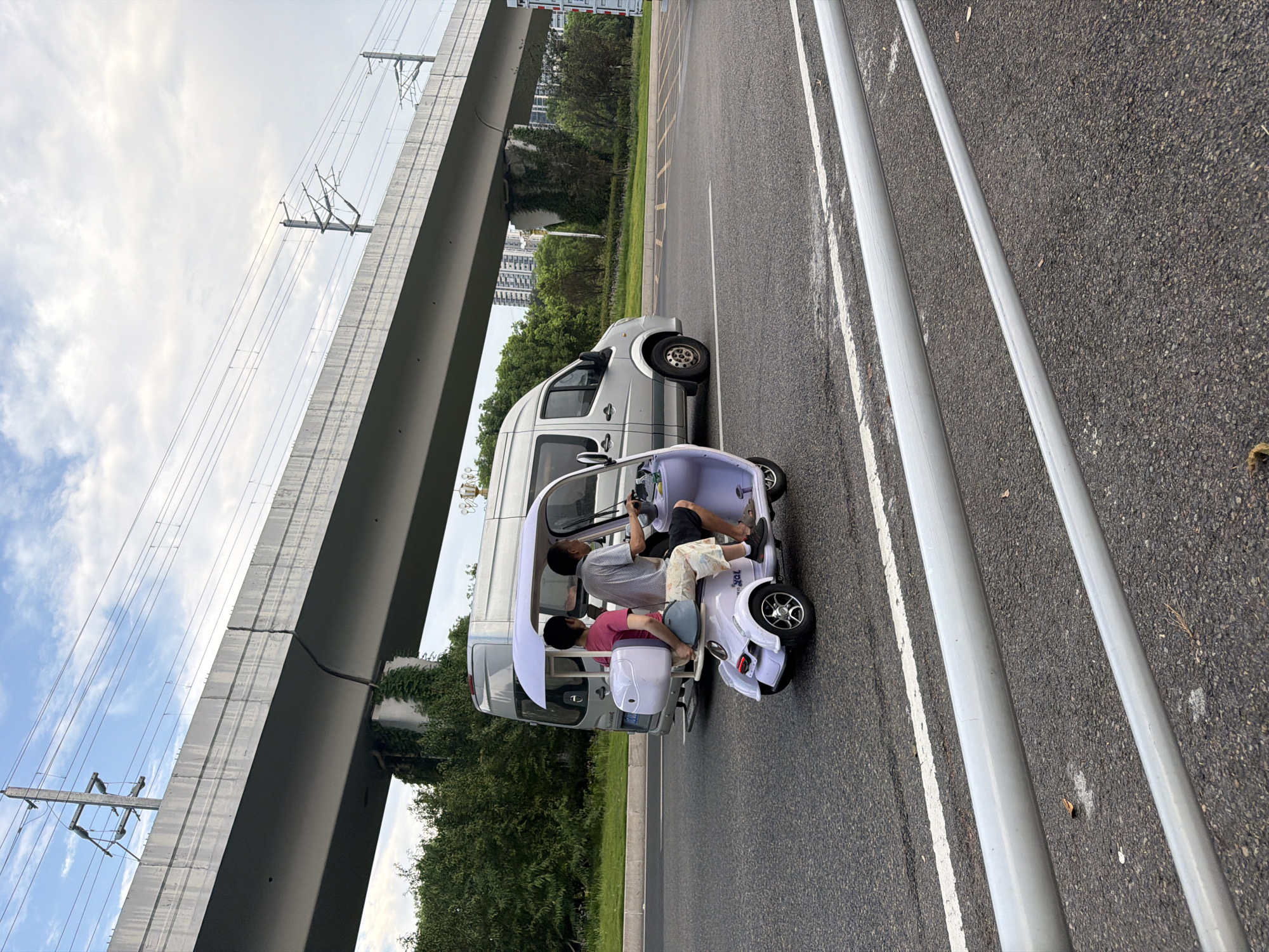 In Jiangjin, China, an overhead overpass with electrical wires and a crack running down its side looms over a road. On the road, two individuals are seated in a light purple, three-wheeled electric vehicle. The driver, a male wearing a checkered shirt and patterned shorts, operates the vehicle. Behind him, a female in a pink shirt and checkered shorts sits. A light gray van is positioned behind them on the road. The scene is set with green foliage and trees lining the side of the road, with tall buildings visible in the distance. The sky is partly cloudy.