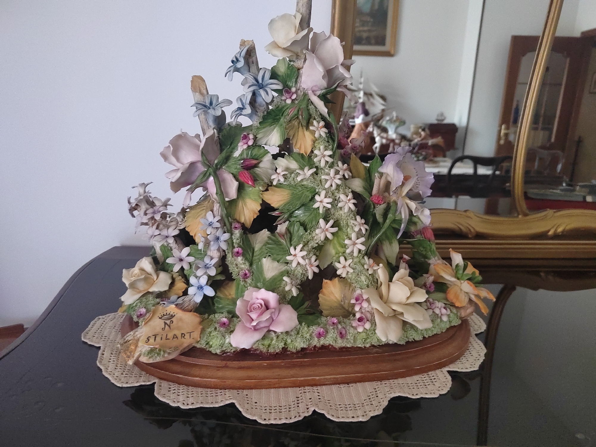 The image depicts a detailed porcelain figurine of a floral arrangement, predominantly featuring pink, blue, and cream-colored flowers, with green foliage. The figurine is placed on a wooden base, which is in turn situated on a crocheted lace doily. The doily rests on a dark, reflective surface, likely a table. In the background, a large, ornate gold mirror reflects a room with furniture, including a wooden cabinet and chairs, suggesting an indoor setting. The overall impression is one of an ornamental display piece, possibly within a home or a shop. The specific location of Porto Recanati, Italy, is not visually identifiable from the provided image.