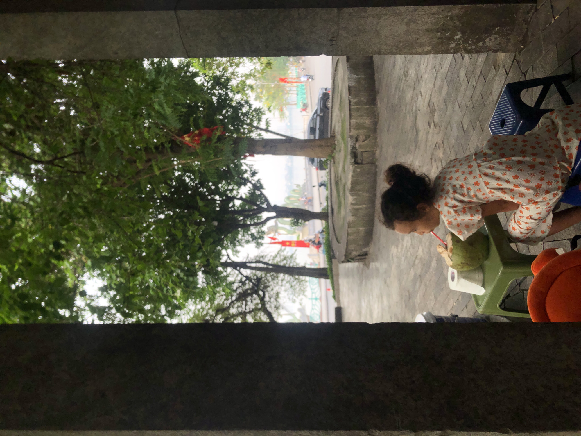 From a narrow perspective, a person is seen sitting, sipping from a coconut through a straw. The individual has dark hair styled in a bun. They are wearing a light-colored, floral-patterned top. The coconut rests on a small, green, child-sized chair or stool. A portion of an orange backpack is visible to the right. In the background, blurred by the perspective, is a street scene featuring greenery, trees, parked vehicles, and Vietnamese flags. The vantage point appears to be inside a structure, with framing pillars visible at the top and bottom of the image. Given the flags and the general environment, the location is likely in the city of Hà Nội, Vietnam.