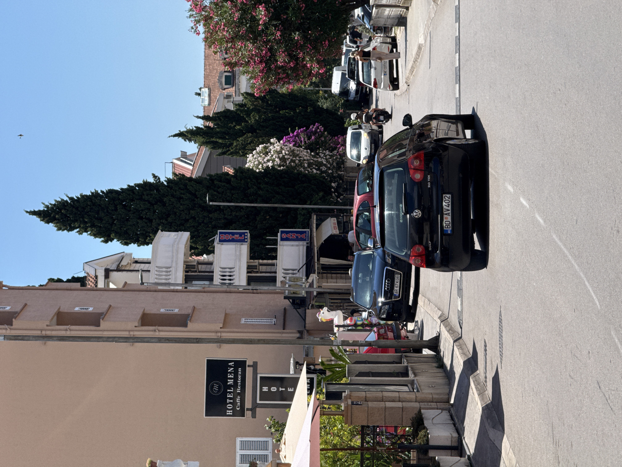 The image shows a street scene in Budva, Montenegro. The street is paved and lined with buildings and trees. Several cars are parked and driving on the street. A black Volkswagen Golf is in the foreground, with its rear facing the camera. An Audi Q5 is parked to its left. A woman in a light-colored dress is walking on the sidewalk to the right. In the background, there are more cars, buildings with balconies, and lush green trees, some of which are adorned with pink flowers. A sign for "HOTEL MENA" is visible on the left side of the street. The sky is clear and blue.