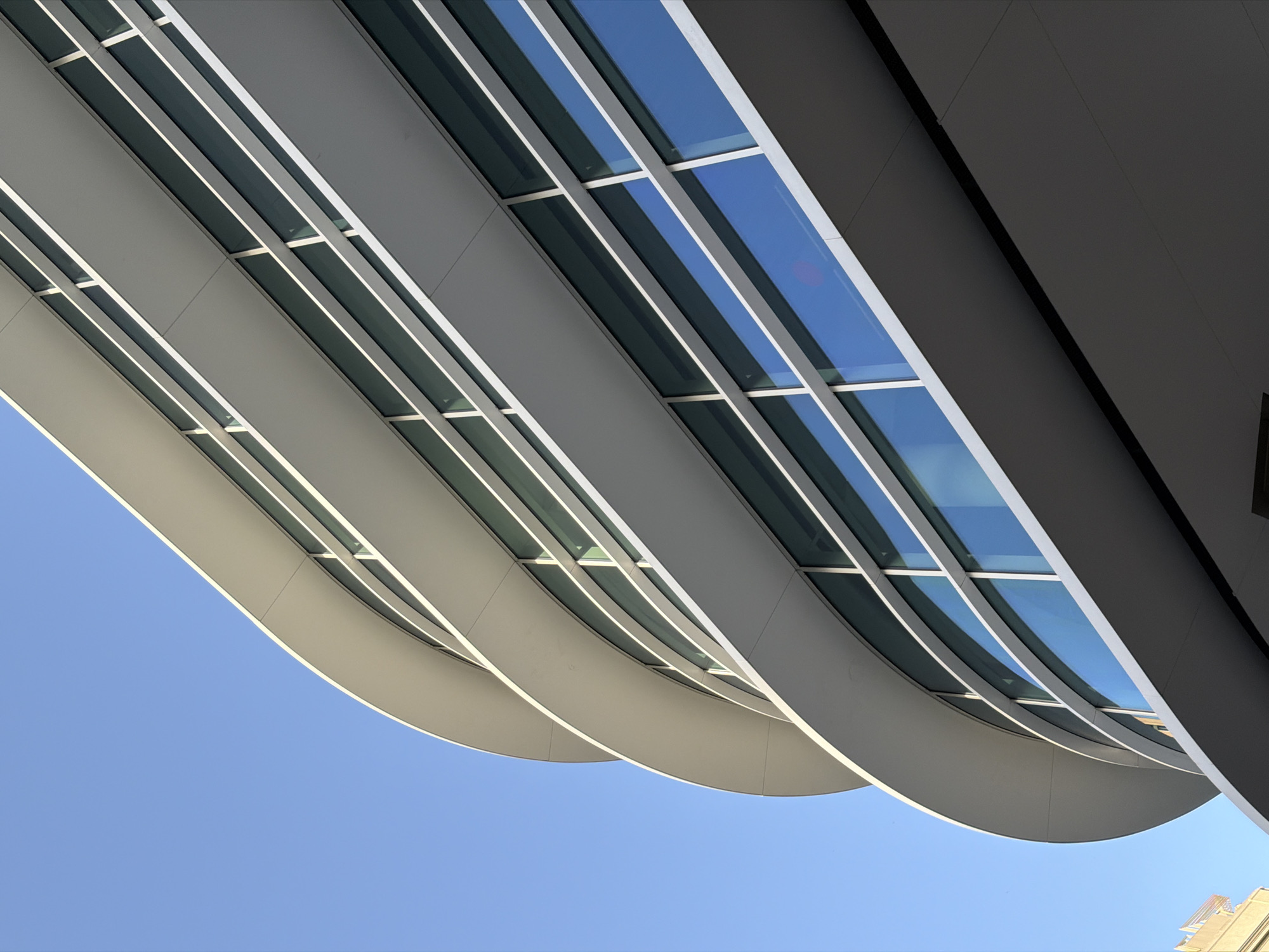 This is a close-up, low-angle, eye-level shot of a modern building in Barcelona, Spain. The building features a curved, white facade with multiple horizontal levels of windows. The windows are a dark, reflective blue, mirroring the clear, bright blue sky above. The building's architecture is sleek and contemporary, with smooth, rounded edges and clean lines. The lower part of the image shows a darker, shaded section of the building, suggesting it overhangs another structure or street. There are no people visible in the image. The overall impression is one of architectural modernity against a pristine sky.